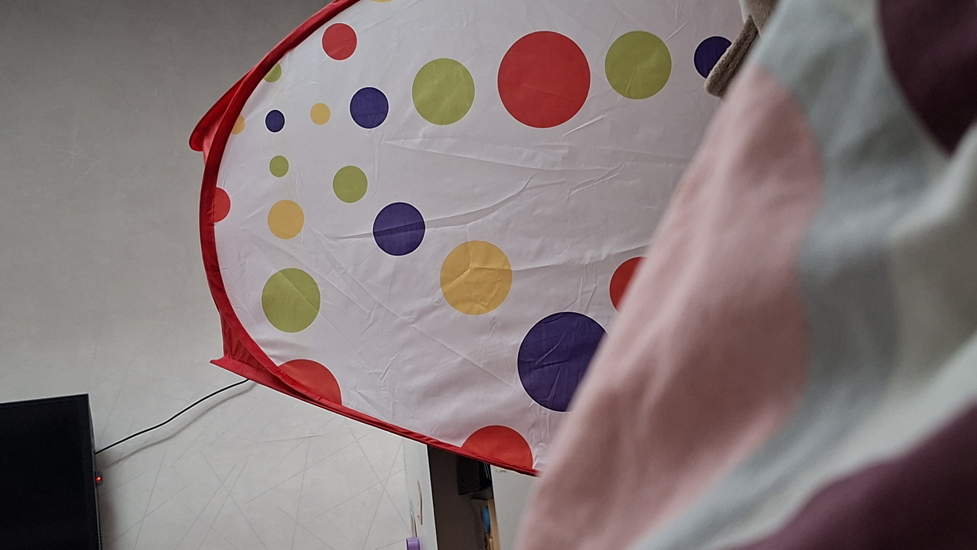 A close-up shot shows a child's pop-up tent with colorful polka dots, partially obscured by a pink and white striped object (likely bedding or clothing). The tent has a red trim. A television is visible in the lower left corner. The wall behind the tent is light-colored with a subtle pattern. The location is not discernible from the image.
