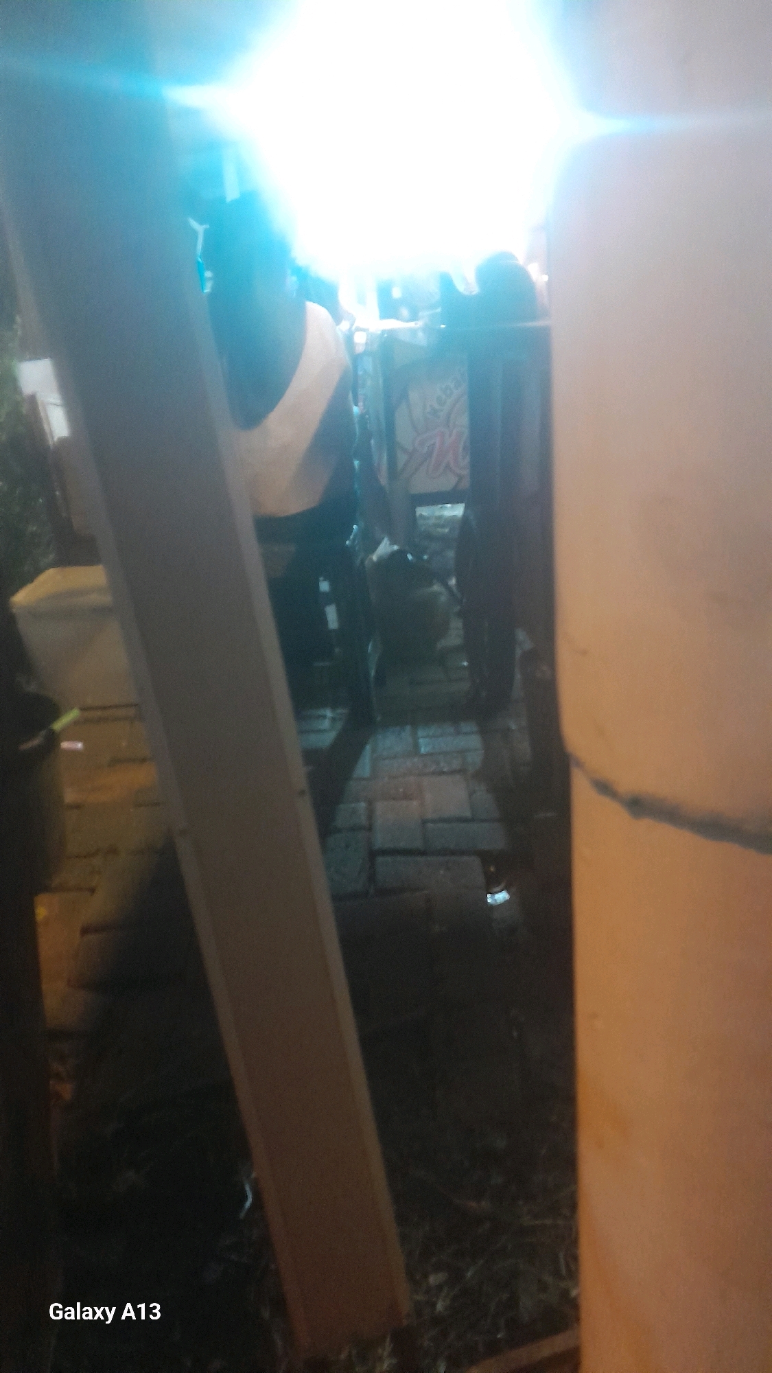 The image depicts an outdoor scene at night, likely in a public space in Ngembalrejo, Indonesia, given the presence of a "Galaxy A13" watermark in the lower left corner, indicating it was taken with a Samsung Galaxy A13 phone. A brightly lit area, possibly a street or a market, is visible in the background, with a strong light source creating a lens flare effect at the top of the image. In the foreground, a person, whose back is turned to the camera, is wearing a light-colored top and dark pants. They appear to be standing near a cart or stall, with a sign that has some lettering on it, partially obscured by the light. The ground is paved with rectangular stones, and there are some plants and debris scattered around. The overall atmosphere suggests a lively evening setting, possibly a food stall or a gathering.