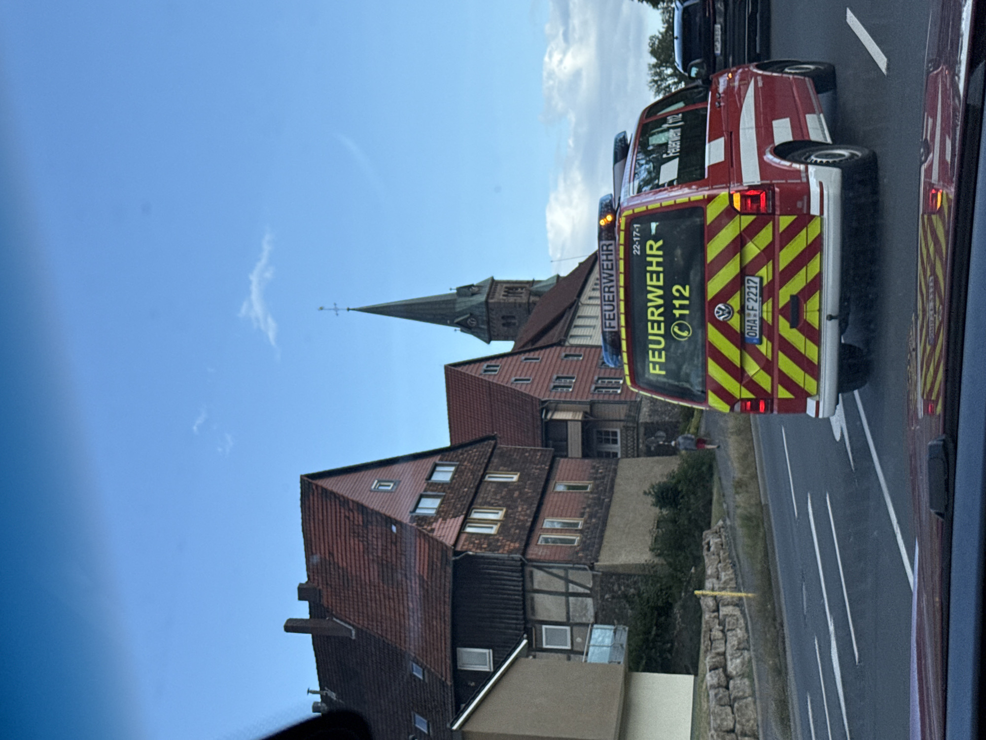 The image depicts a red and yellow checkered emergency vehicle, identified as "FEUERWEHR" (fire department) and displaying the number "112", driving on a road. The vehicle has emergency lights on its roof. In the background, traditional German-style buildings with red tile roofs and exposed timber framing are visible. A church steeple can be seen rising above the buildings. The scene appears to be in Herzberg, Germany, based on the architectural style and the presence of a fire department vehicle. The sky is a clear blue with a few wispy clouds. The photo is taken from a slightly elevated perspective, possibly from inside another vehicle.