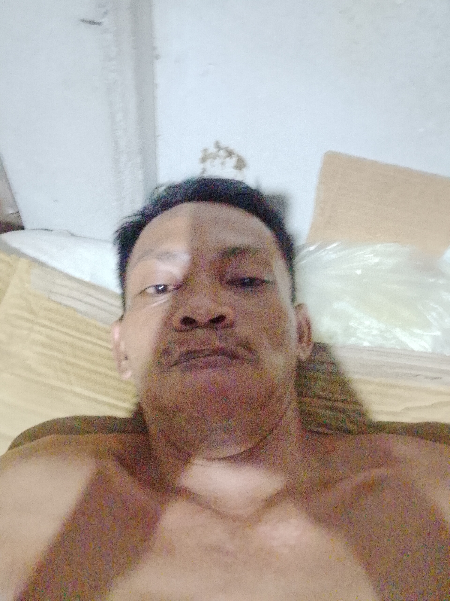 A close-up, low-angle photograph shows a shirtless man lying on a bed in a room in Bukit Ketok, Indonesia. The man has dark hair and is looking directly at the camera. Behind him is a white pillow and a cardboard box. The room's ceiling is white.