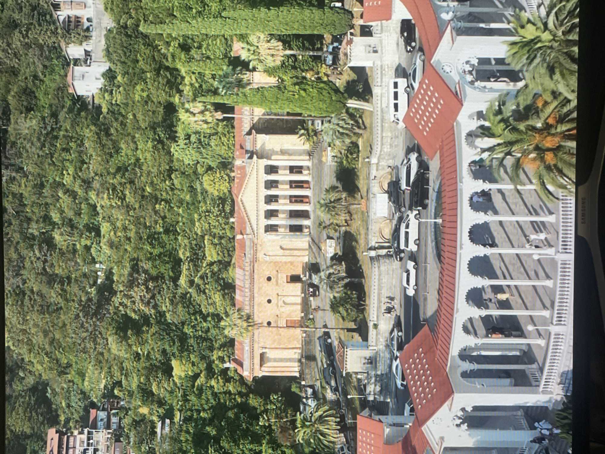 The image shows a wide shot of a large, classical-style building with a red-tiled roof and a colonnade, situated on a hillside covered with lush green trees. In the foreground, there is a white, open-air structure with a red-tiled roof, also featuring a colonnade and a white railing. Several cars, including vans and sedans, are parked on a road in front of the main building and on a street in front of the foreground structure. A few people can be seen walking around the foreground structure and on the steps leading up to the main building. The main building is likely the Tskneti Guesthouse in Abkhazia, located in the city of Sokhumi. The overall impression is one of a scenic, historic location, possibly a resort or a public building, surrounded by nature.