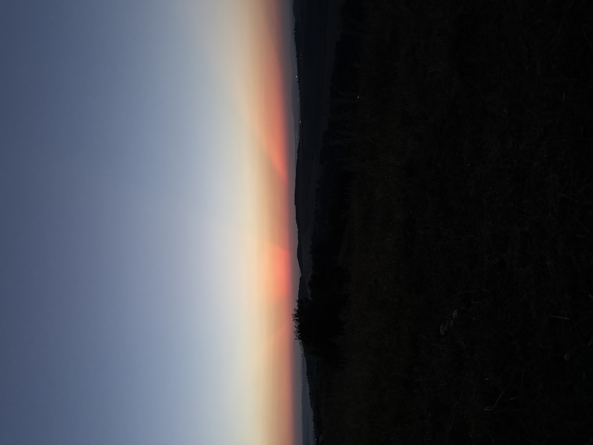 The image depicts a twilight landscape with a gradient sky transitioning from deep blue at the top to a soft orange and pink near the horizon. In the distance, rolling hills are silhouetted against the fading light. A dark mass of vegetation, possibly a tree or a dense bush, stands prominently in the mid-ground. The foreground is filled with dark, out-of-focus vegetation, suggesting it is a field or grassy area. The overall impression is of a serene, natural setting at dusk. The location is described as Unknown, Unknown. There are no people or discernible actions in the image.