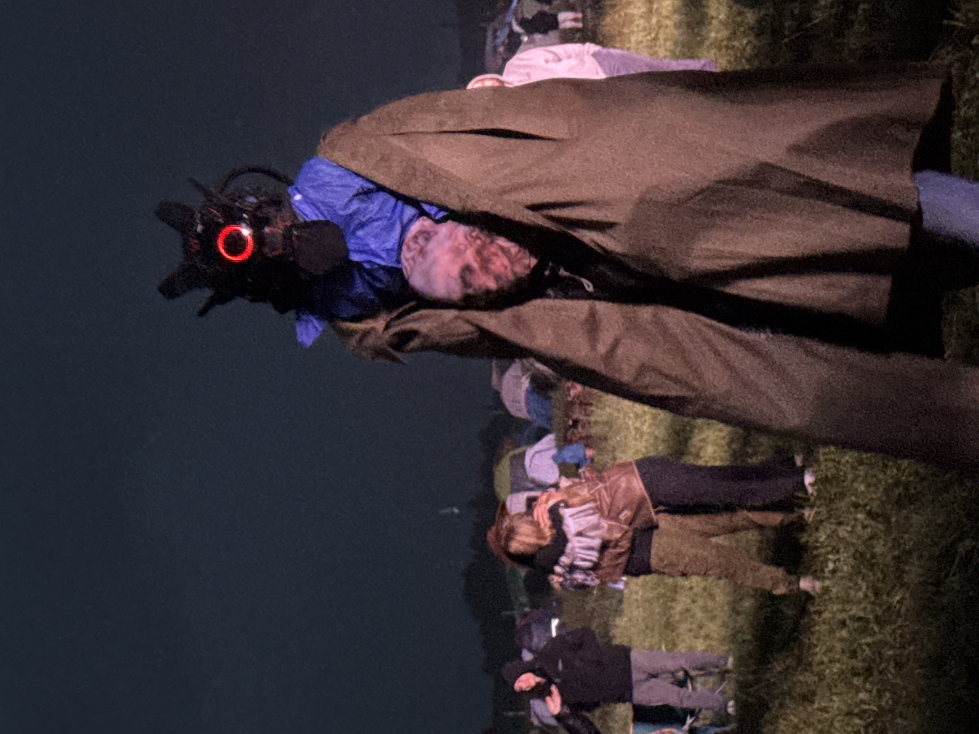 The image depicts an outdoor night scene in a grassy field in Большая Кивара, Russia. The central figure is a tall individual partially visible on the right, enveloped in a dark brown, floor-length cloak. Beneath the cloak, a blue garment covers their upper body, from which a dark, intricate mask with spiky protrusions and a prominent, glowing red circular light emanates. Below the mask, a pale, disfigured face-like prop with pinkish-red tones is visible. The figure's lower legs, clad in blue pants, are visible beneath the cloak. To the left, two individuals are embracing; one wears a patterned top and light brown pants, the other a brown jacket and dark pants. Further left, a person in a dark hooded sweatshirt and light grey pants stands, gesturing with their right hand. In the background, numerous tents of various colors (grey, blue, green, purple) are dispersed across the grassy terrain. The sky is dark, indicating night, with a faint light source visible in the distance above a dark treeline on the horizon.