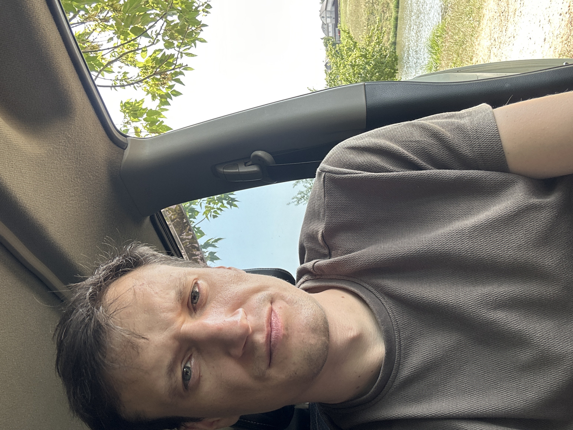 The image shows a selfie taken from inside a car. The driver, a white male with short, dark hair, is looking at the camera. He is wearing a dark grey t-shirt. Through the car window, a rural landscape is visible, featuring a body of water, green fields, and some houses in the distance. Lush green trees with sunlight filtering through their leaves are also present. The interior of the car, including the car ceiling and a part of the car door, is also captured in the frame. The scene appears to be set in a sunny, outdoor environment.