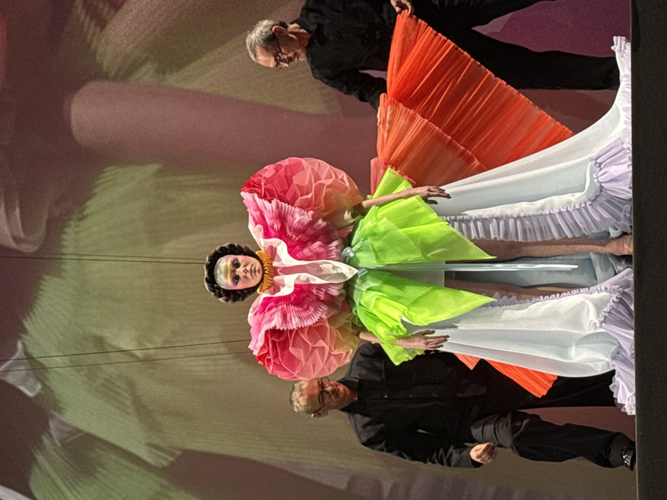 A vertical view shows a puppet-like figure in an elaborate costume reminiscent of a flower. The dress features layers of colors—pink, green, orange, white, and lavender—constructed from pleated fabric. Two men in black attire flank the figure. One holds the orange part of the costume. The entire scene appears to be set against a softly colored backdrop of pastel shades, possibly a stage projection. The figure appears to be suspended.