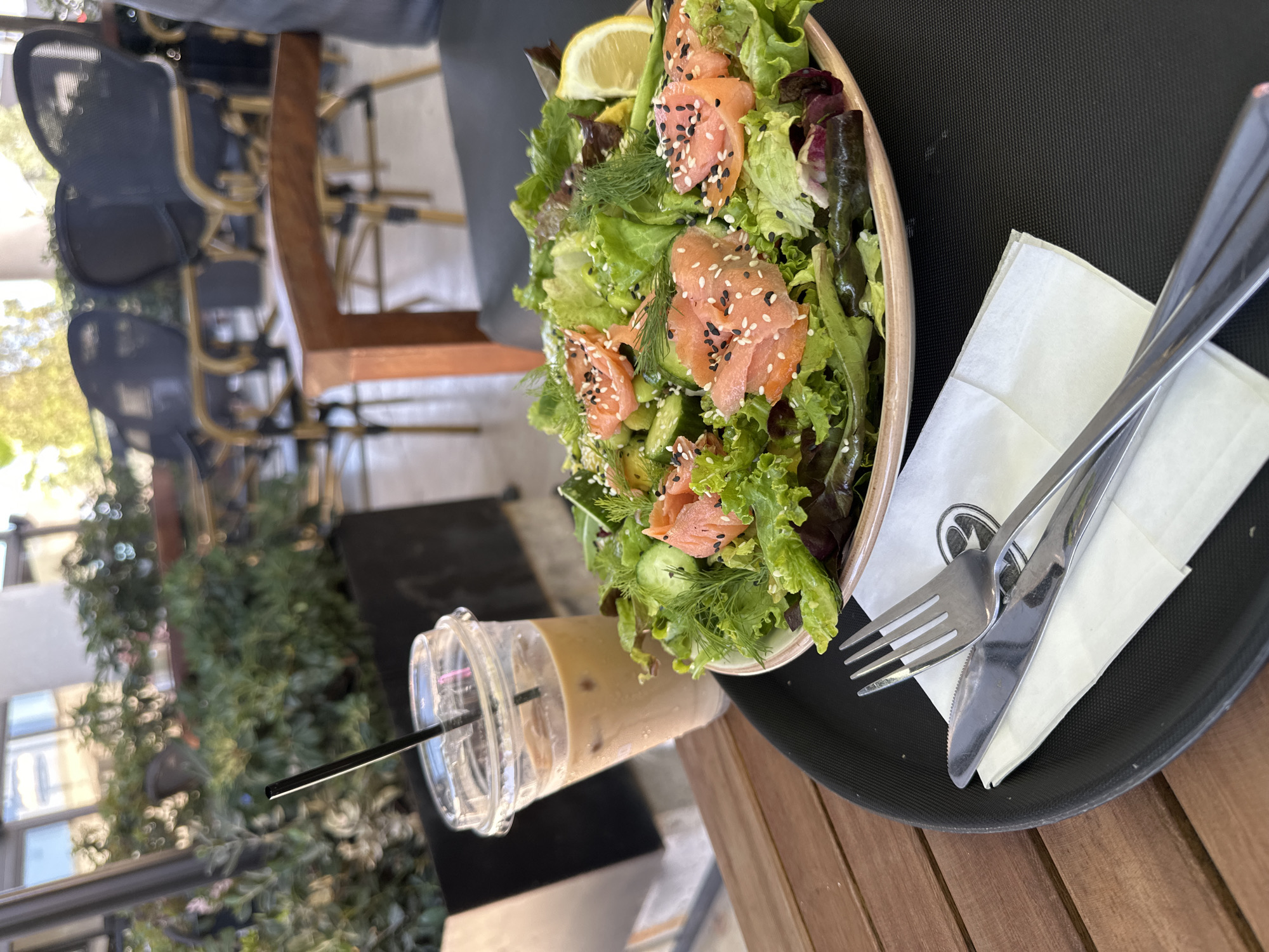 A salad and a beverage sit on a wooden table. The salad is served in a light-colored bowl and is composed of mixed greens, sliced salmon, cucumbers, dill, and sesame seeds. A lemon wedge is perched on the side of the bowl. To the left of the salad is a clear plastic cup filled with an iced coffee beverage with a black straw. A fork and knife are positioned on a white napkin to the left of the salad. The setting appears to be an outdoor seating area, likely a cafe or restaurant, with blurred background elements of chairs and greenery, suggesting a location in Paphos Municipality, Cyprus. The lighting suggests it is daytime.
