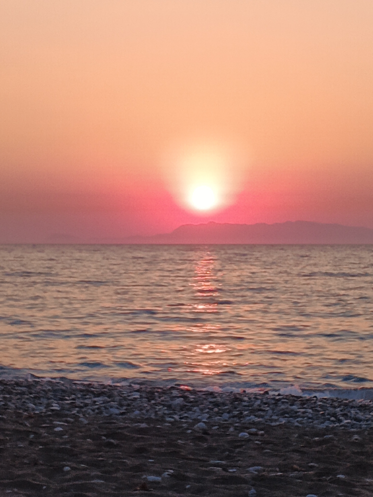 A vertical, close-up shot captures a serene beach scene at sunset. The sun, a brilliant orb, is partially obscured by the horizon, casting a warm, orange glow across the sky. The water, calm and reflective, shimmers with the sun's rays, creating a mesmerizing pattern of light. The foreground is dominated by a pebbled beach, with dark sand and scattered stones. The distant shoreline is lined with a silhouette of mountains, adding depth and a sense of tranquility to the image. The overall atmosphere is one of peace and natural beauty.