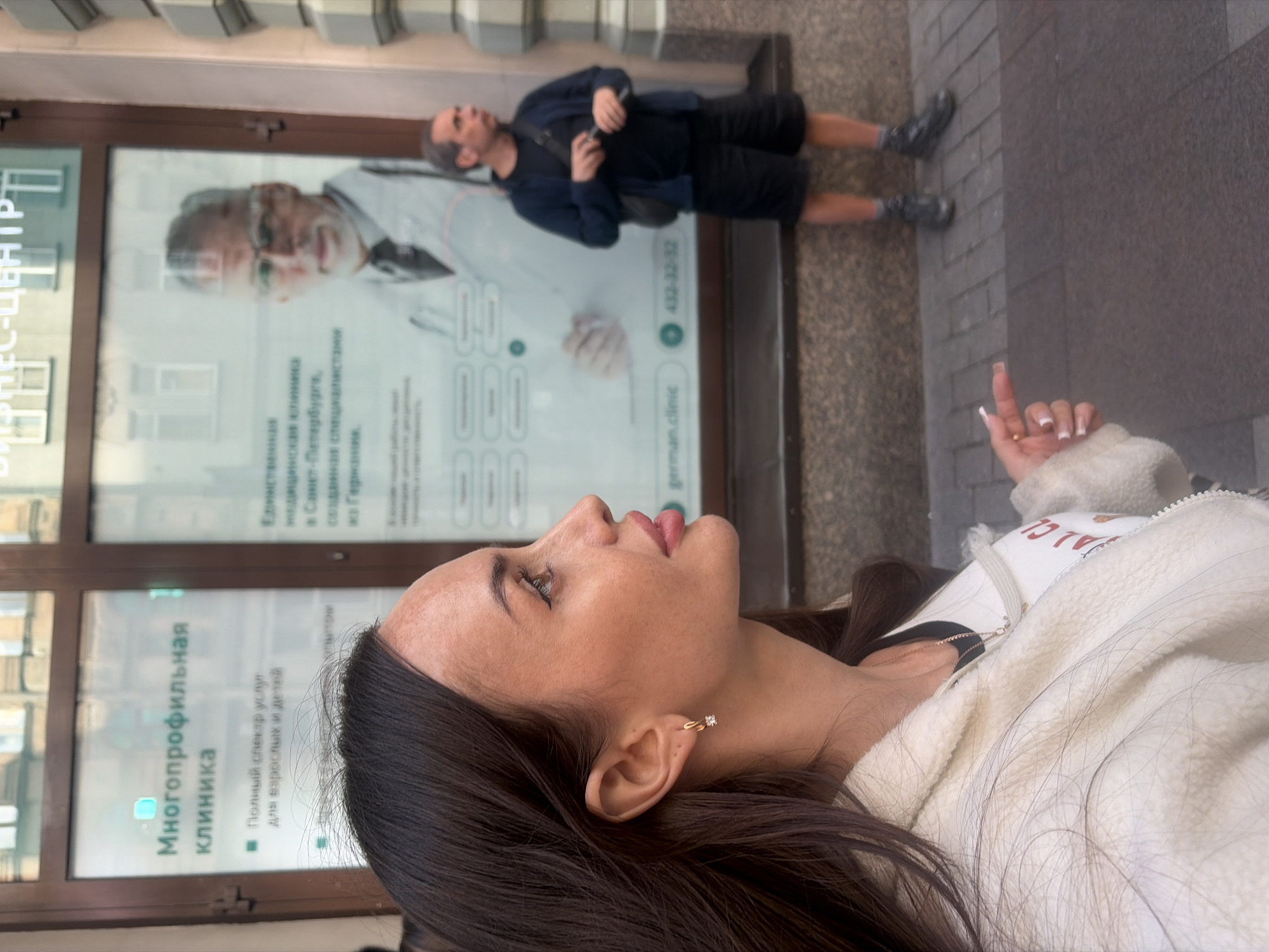 A woman with dark hair, wearing a white fleece jacket and a necklace, is in the foreground of an image. Her head is turned to the right, and she appears to be looking slightly down and to the right. She has an earring in her left ear. Behind her is a shop window with advertisements. One advertisement features a blurred image of a man in a doctor's coat and glasses, with text in Russian. Another advertisement has text in Russian, including "Многопрофильная клиника" (Multidisciplinary clinic). A phone number "432-32-32" and a website "german.clinic" are visible. In the background, a man in a dark t-shirt, blue jacket, and black shorts is standing, looking to the right. The scene appears to be on a street in Saint Petersburg, Russia, indicated by the text on the advertisements. The ground is paved with rectangular stones.
