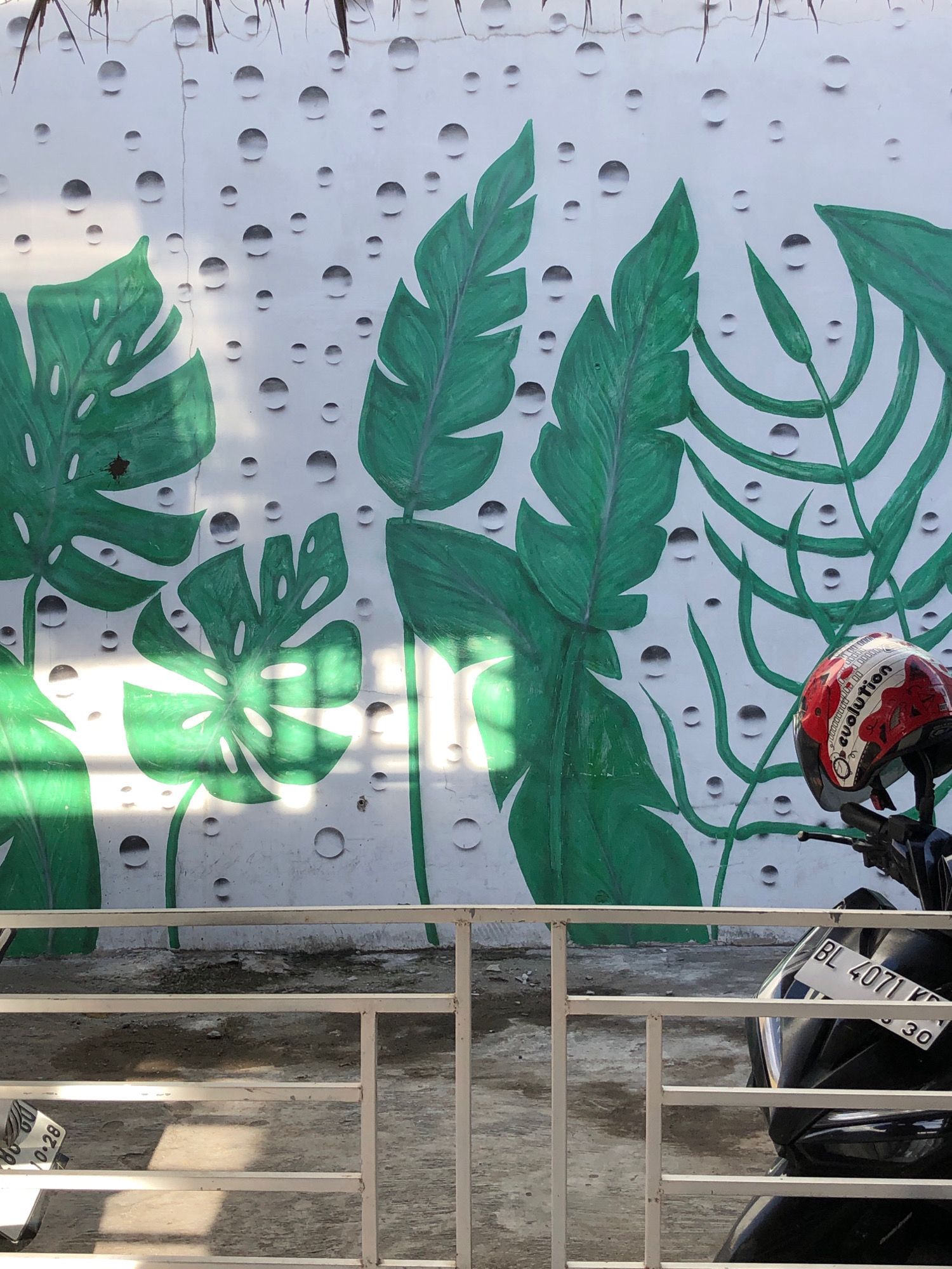 In Uteungeulinggang, Indonesia, a street scene unfolds with a mural adorning a white wall. The mural depicts large green tropical leaves against a backdrop of painted water droplets. In the foreground, a white metal fence separates the viewer from the wall. To the right, a black motorcycle is parked, its handlebars visible. A red and white helmet with "evolution" written on it rests on the handlebars. A license plate on the motorcycle reads "BL 4071 KP". The ground appears to be paved and somewhat wet, with shadows cast by the fence and possibly sunlight filtering through.
