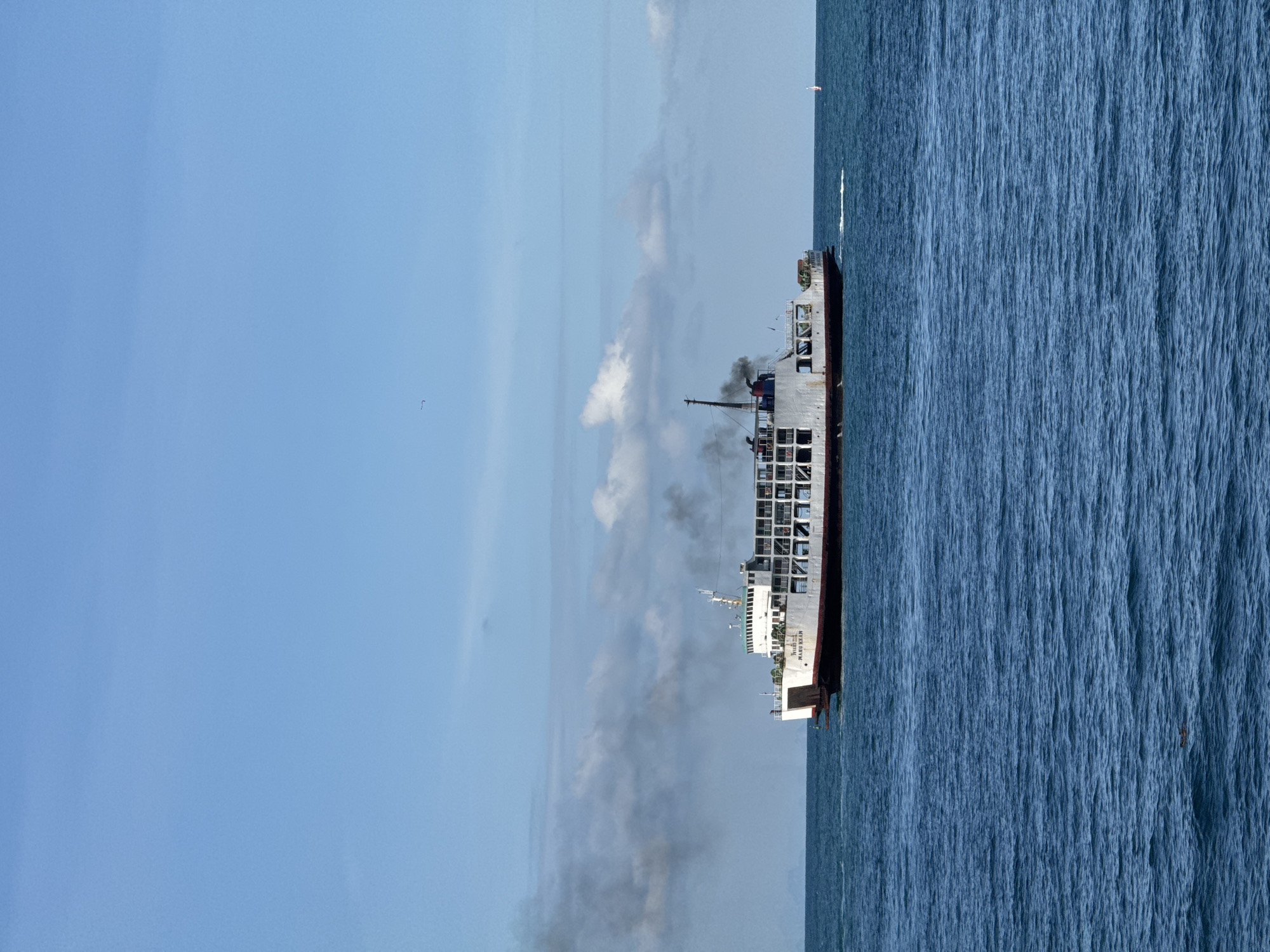 A white ferryboat, identified by the text "MV RORO KELU" on its side, sails on a calm, blue sea under a clear sky with scattered white clouds. The ferryboat emits dark smoke from its smokestack. The vessel appears to be a Ro-Ro (roll-on/roll-off) ferry, designed to carry vehicles. The overall scene suggests a routine journey at sea. The location is specified as Tambon Koh Phangan, Thailand.