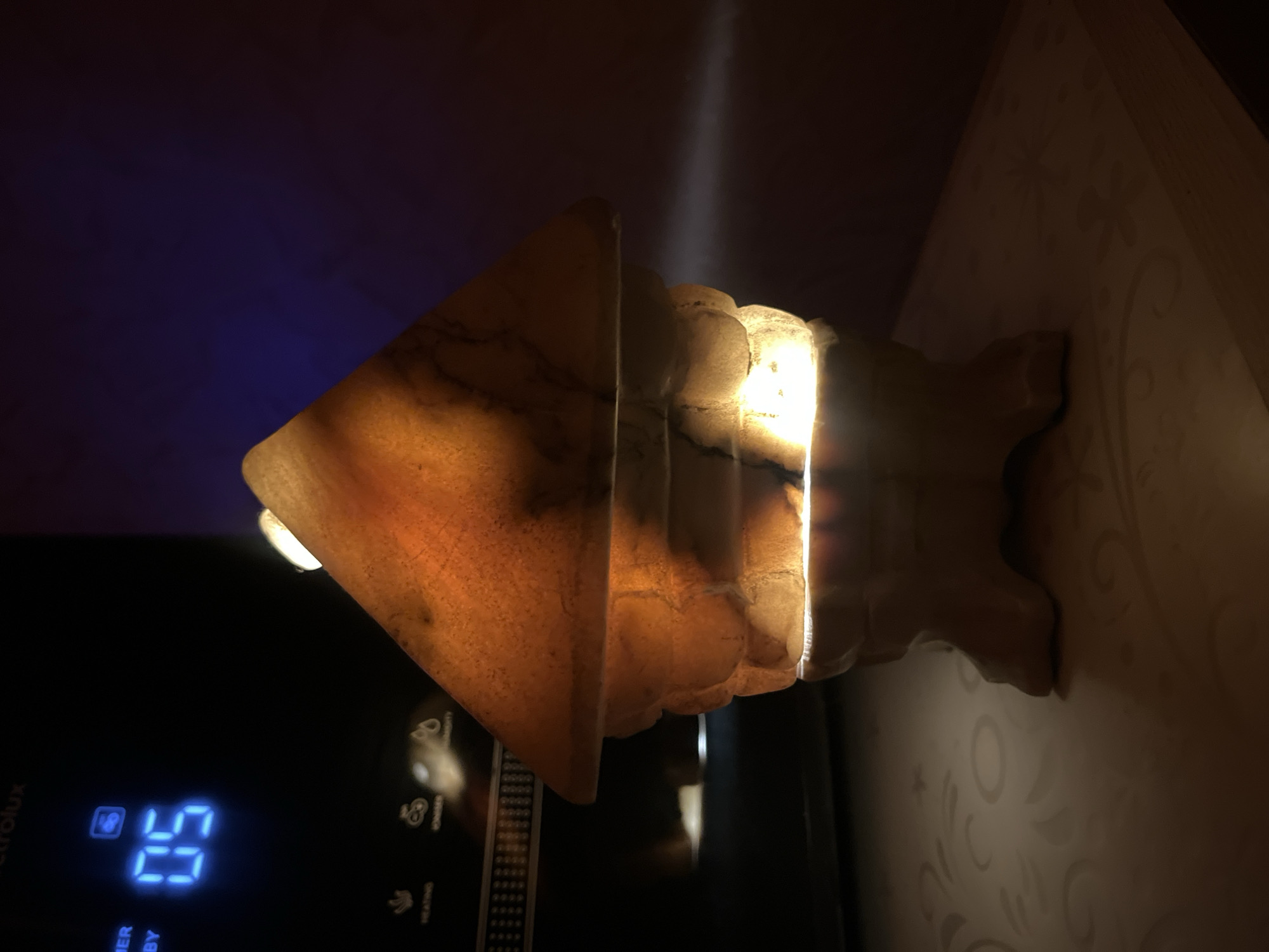 A lit salt lamp shaped like a house is positioned on a patterned surface. The lamp, made of translucent, amber-colored material with dark veins, emits a warm glow from within. Behind it, a dark electronic device with a blue digital display showing "05" is visible. The background is dark with a subtle blue light gradient. The overall scene is dimly lit, with a beam of light emanating from the lamp to the right. The location is identified as Kyiv, Ukraine.