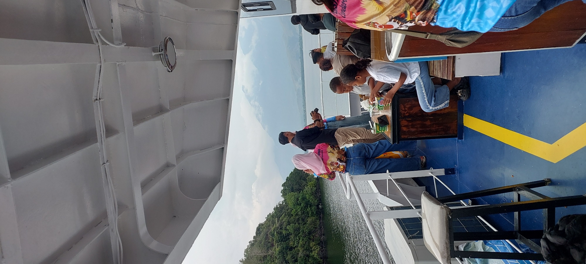 The image shows a view from the deck of a ferry on a cloudy day, likely in Hobong, Indonesia. A body of water, possibly a lake or sea, is visible in the background with lush green hills and mountains under a gray sky. Several people are on the deck, some standing and others seated. A woman in a pink hijab and a colorful jacket, along with a man in a black shirt, are standing near the railing, facing the water. A group of children and adults are seated at a table, with some of them appearing to eat or drink. There are also chairs and other seating arrangements on the deck, as well as some luggage. The deck is painted blue with a yellow line marking a pathway. The overall scene suggests a relaxed outing or travel experience on the water.