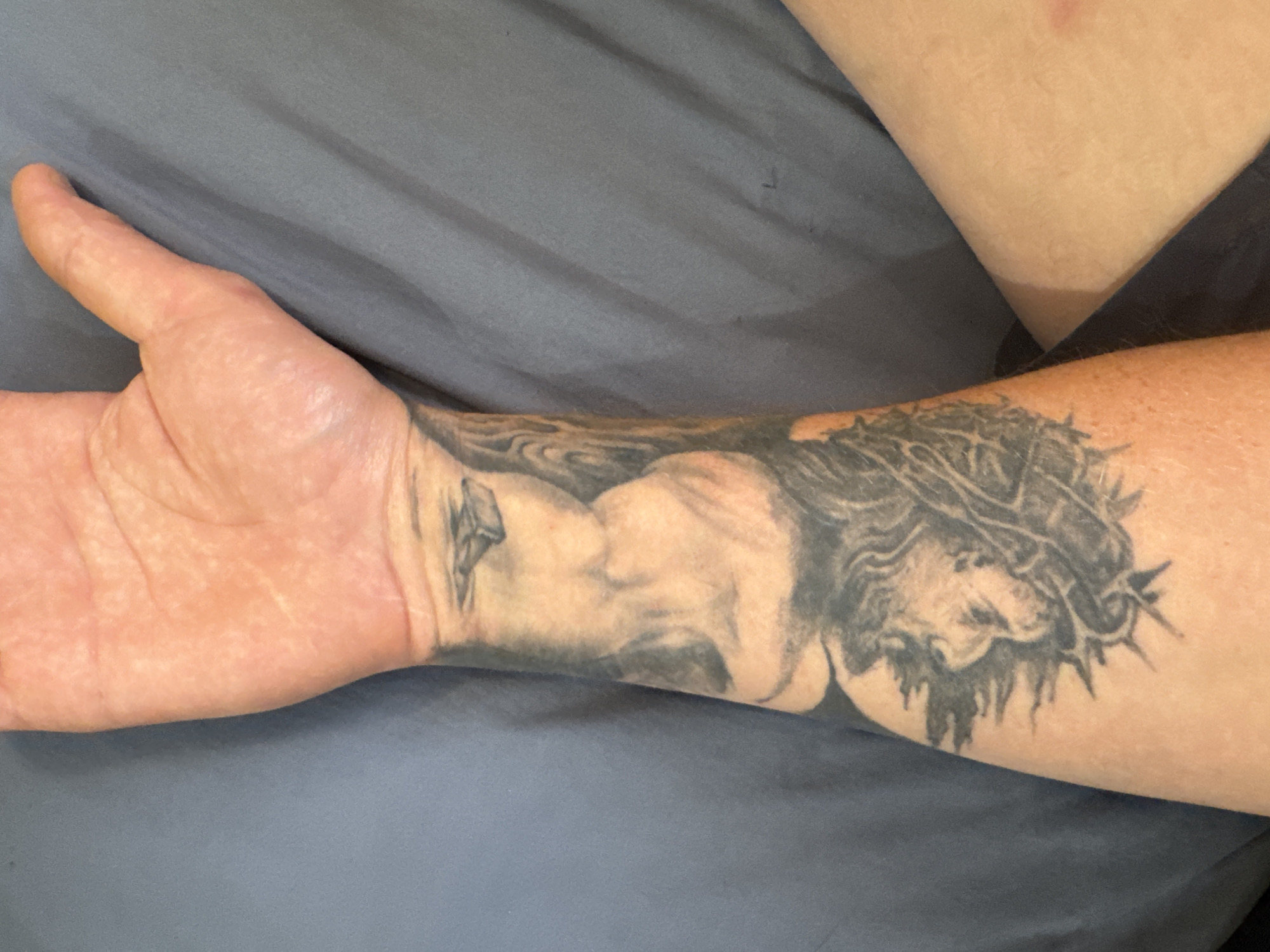 A person's forearm, displaying a black and grey tattoo, rests on a dark blue fabric surface. The tattoo appears to depict a lion's head with a spiky crown, and possibly other figures or designs further up the arm. The skin on the arm is lightly tanned and shows some texture. The fabric beneath is wrinkled, indicating it is likely a sheet or blanket. The image is tightly cropped, focusing on the forearm and a portion of the hand, with no indication of the city of Protasovo, Russia.