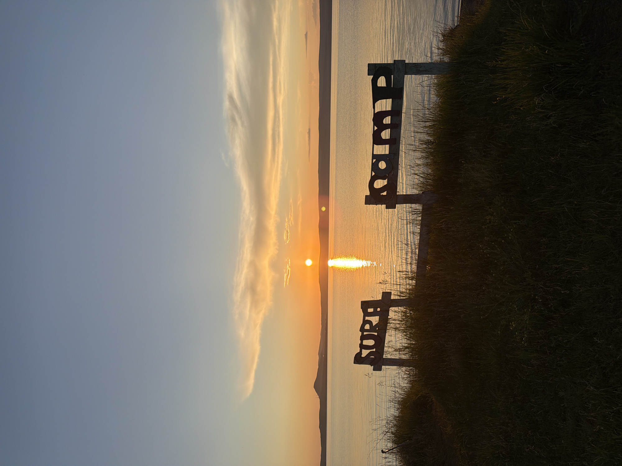 An outdoor, horizontal, medium shot captures a sunset over a body of water in Russia, at an unknown location. The sun is setting on the horizon, casting a warm orange glow across the sky and reflecting on the calm water. The sky transitions from a pale blue at the top to a soft orange and yellow near the horizon, with wispy clouds. In the foreground, two wooden signs are visible, partially obscured by tall grass. The sign on the left appears to read "SURF," and the sign on the right reads "CAMP." The overall scene is tranquil, with the setting sun creating a peaceful atmosphere.