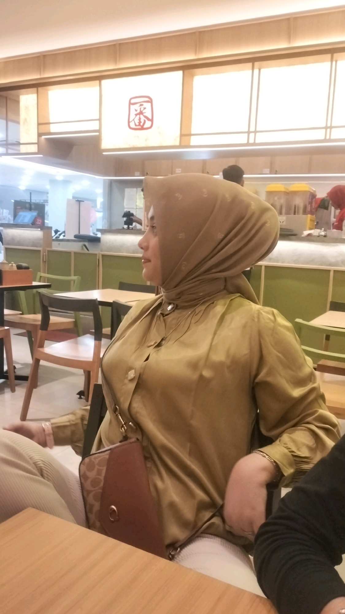A woman, wearing a tan hijab and a tan blouse, is seated at a table in a restaurant. She is looking to her right. A brown purse with a tan pattern is hanging from her shoulder. The restaurant appears to have a modern, minimalist design with wooden tables and chairs, and illuminated panels on the ceiling with a red symbol. The background shows other tables and chairs, as well as kitchen equipment. The woman is sitting in the foreground, with the rest of the restaurant visible behind her. It is daytime.