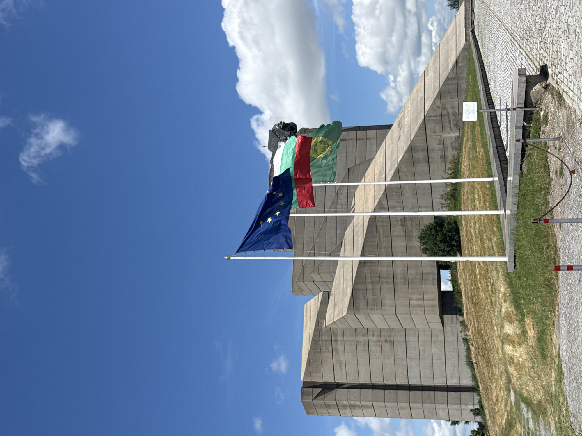 The image captures the Monument to the Founding of Bulgaria in Shumen, Bulgaria. The monumental concrete structure, characterized by its angular and imposing design, dominates the background under a bright blue sky with scattered white clouds. In the foreground, three flags fly from white poles: the European Union flag, the Bulgarian flag, and another flag that appears to be a historical Bulgarian banner with a lion emblem. The flags are positioned on a pathway leading towards the monument, with dry, yellowed grass and some green patches on the hillside surrounding the structure. A paved walkway composed of cobblestones is visible in the lower right portion of the image. A small white sign with a circular emblem is also present near the flags. The scene is devoid of any people.