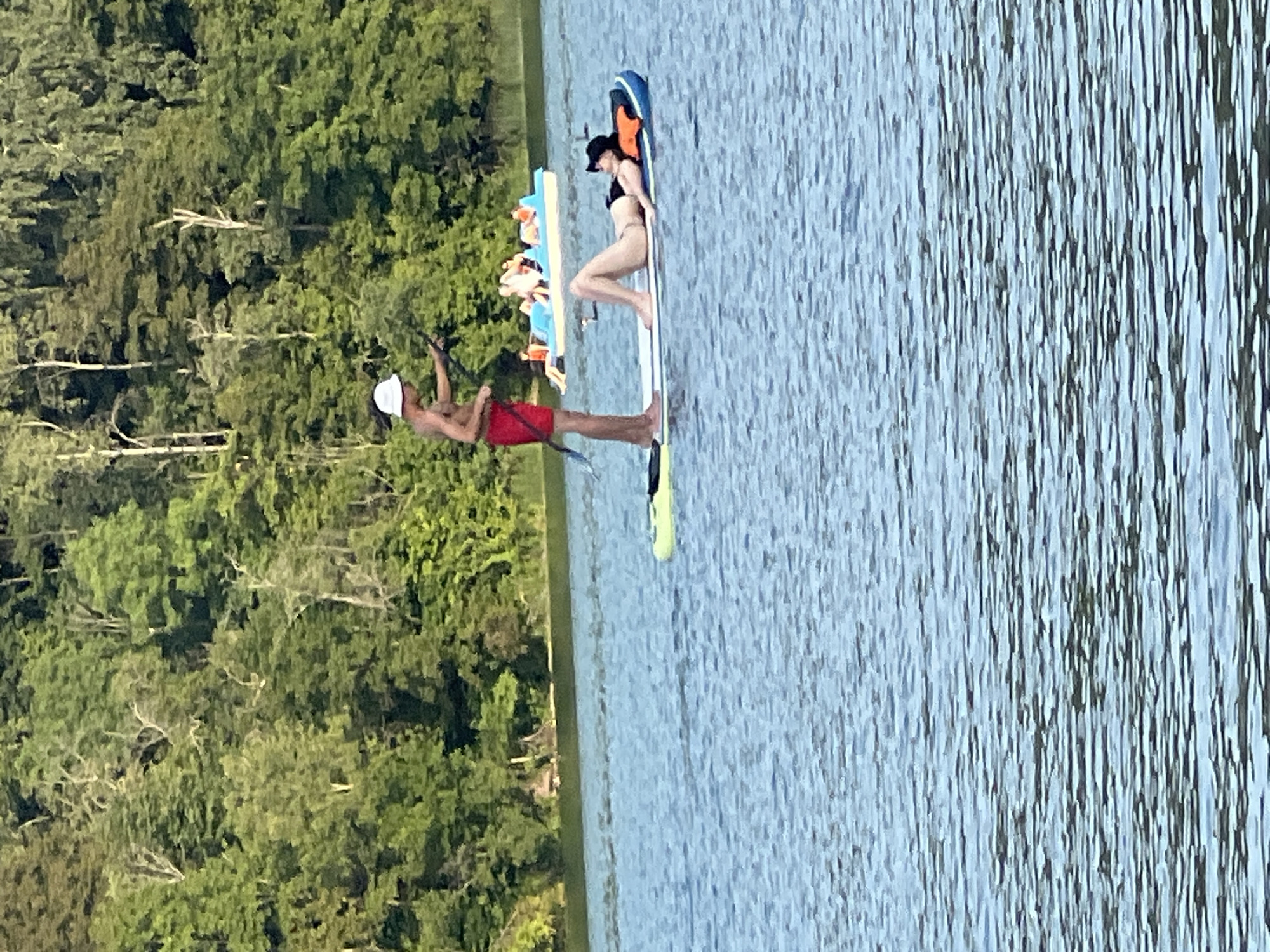 The image captures a serene day on a body of water, likely a lake or river, with a dense forest lining the background. In the foreground, two individuals are present on paddleboards. One individual, wearing red shorts and a white hat, stands on a paddleboard, holding a paddle. To their right, another individual, wearing a black bikini and a black hat, reclines on a paddleboard. In the mid-ground, a small boat with several people is visible, also enjoying the water. The water itself is calm, reflecting the overcast sky. The overall scene suggests a leisurely outdoor activity in a natural setting, possibly a park or recreational area within or near Vilnius, Lithuania, given the common presence of forested lakes and rivers in the city's vicinity.