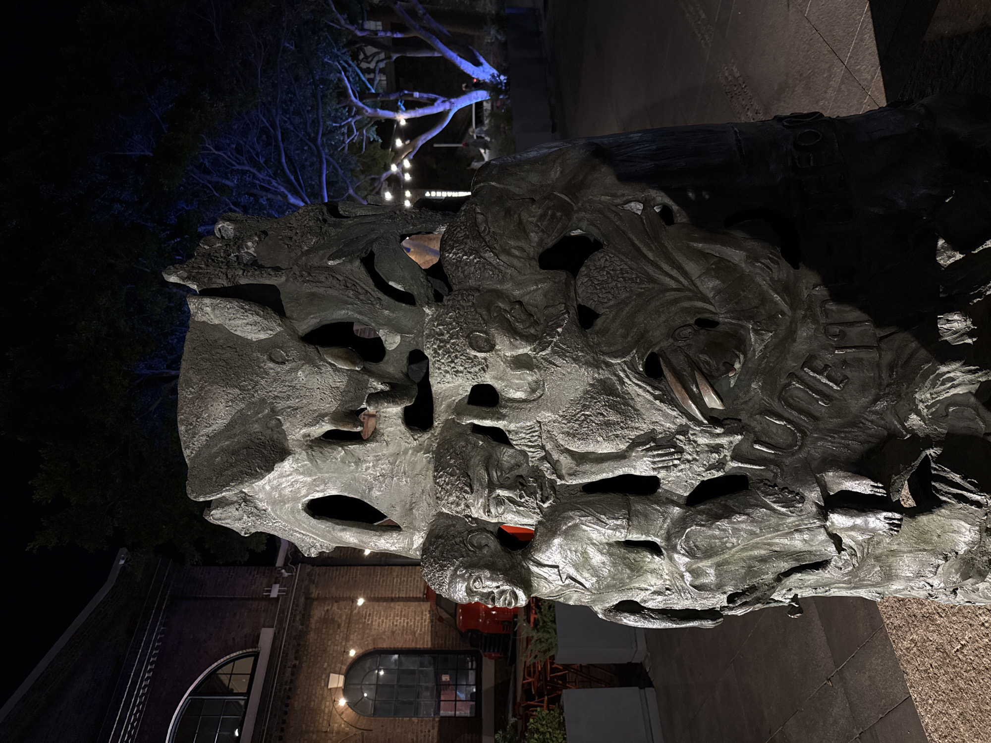 The image shows a sculpture made of what appears to be metal, featuring carved or molded figures. It is situated outdoors at night.
In the background, there is a building with a visible arched window and a round window to the side. The building seems to be constructed of brick. Trees and strung lights can be seen in the distance beyond the building.
The sculpture is lit from below, creating shadows and highlighting its textured surface. A construction lift is visible behind the sculpture. The image is taken at night.
The precise location within Cape Town is impossible to ascertain from the given image alone.