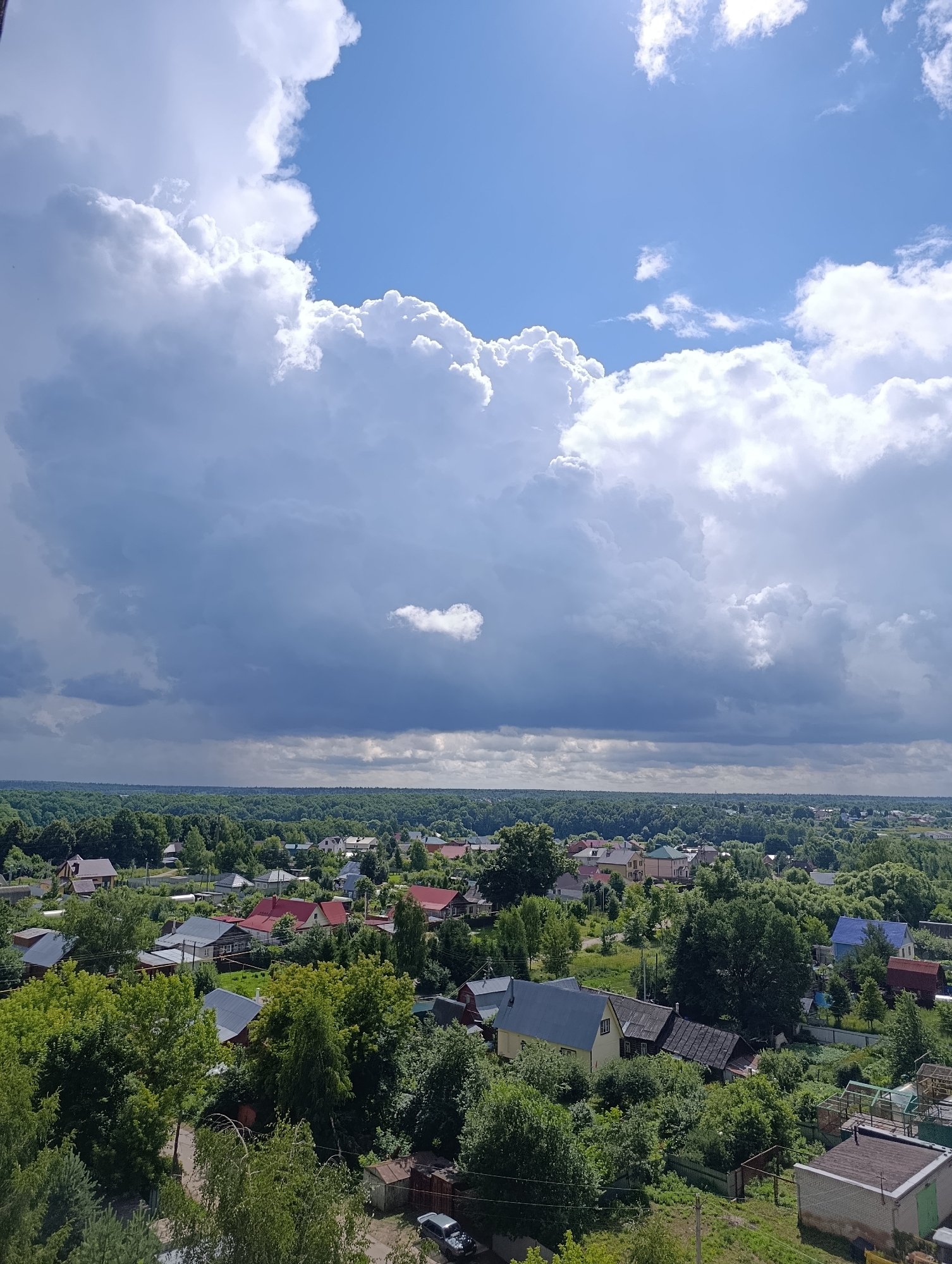 The image presents a panoramic view of a rural settlement, likely on the outskirts of Alexandrov, Russia, under a dramatic sky. The foreground is dominated by lush green trees and foliage, with several houses of various sizes and architectural styles nestled among them. Some houses have red or dark-colored roofs, while others feature lighter hues. A dirt road is visible, with a car parked on it in the lower right portion of the frame.
The mid-ground reveals a denser collection of residential buildings, indicative of a village or a suburban area. The horizon is lined with a dense forest, stretching as far as the eye can see.
The sky is a striking feature, with large, billowing cumulus clouds dominating the upper half of the image. The clouds range from bright white where illuminated by the sun to darker, grayer tones, suggesting a potential for rain or an approaching storm. The overall atmosphere is that of a summer day with dynamic weather conditions. There are no people visible in the scene.