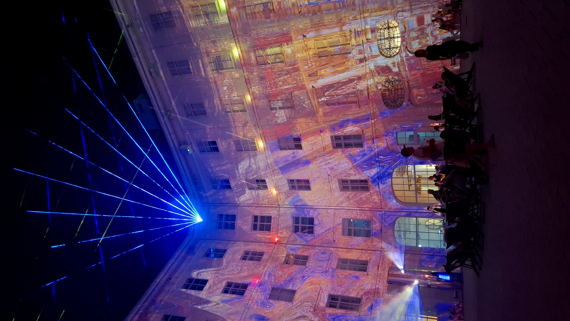 The image captures a night scene at the Volksgarten in Vienna, Austria, where an elaborate light show is projected onto the façade of a building. A powerful blue laser emanates from a central point, fanning out across the dark sky in a radial pattern, interspersed with thinner green laser lines. The building itself is illuminated with a vibrant, abstract projection featuring swirling patterns of pink, orange, and purple, creating an illusionistic marbling effect. Numerous windows on the building are visible, some appearing dark while others reflect the projected light.
In the foreground, the courtyard area is occupied by an audience seated in dark deck chairs, facing the illuminated building. Several individuals are visible, some standing and others seated, observing the spectacle. A projector beam, colored blue and white, is visible on the left side of the courtyard, casting light onto the ground and surrounding area. The ground is paved with a dark, textured material. The overall atmosphere suggests an outdoor event or festival, with the dramatic light display being the central focus.