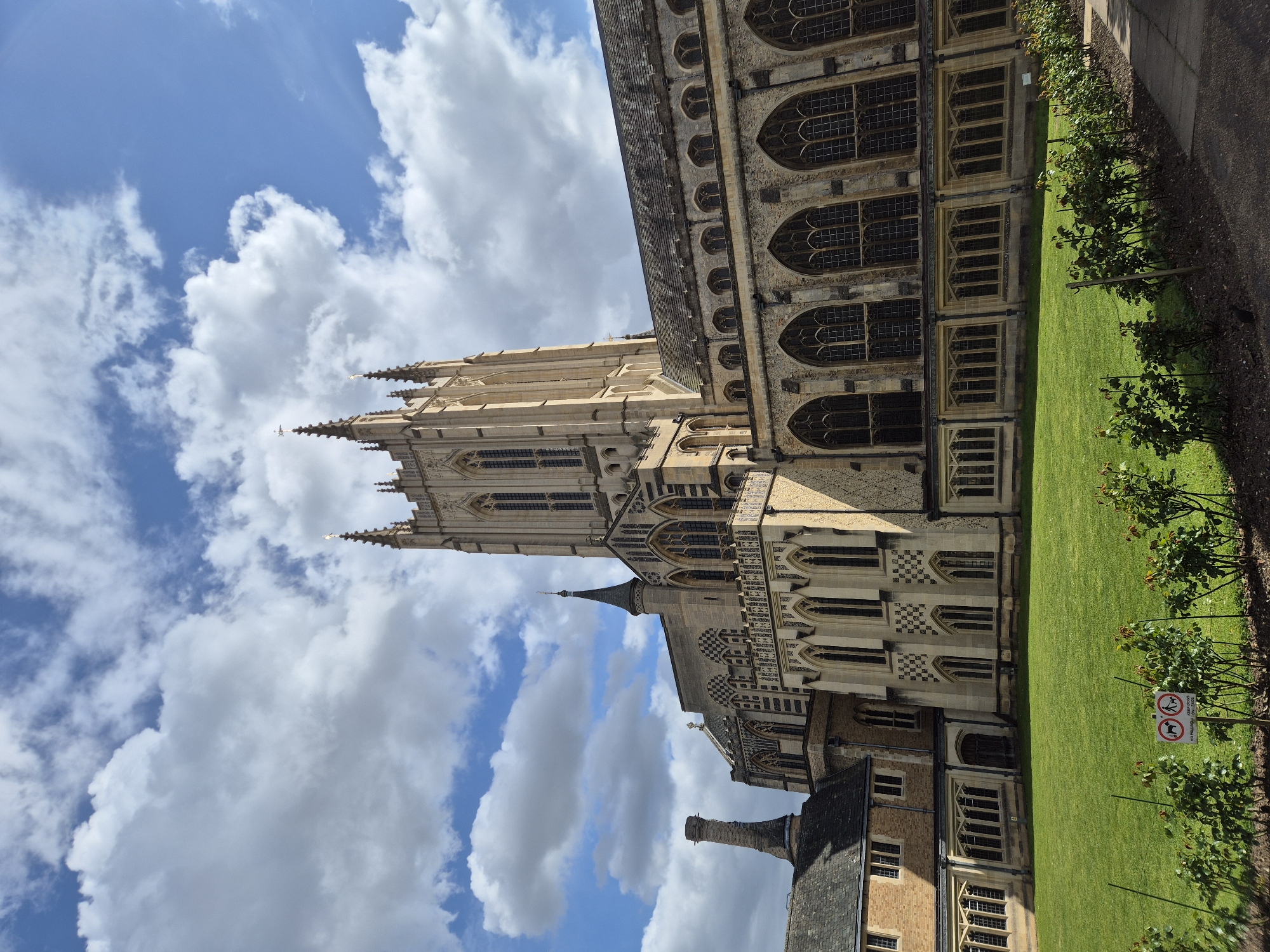 The image captures an upward view of the historic St Edmundsbury Cathedral located in West Suffolk, United Kingdom, showcasing its architectural grandeur against a partly cloudy sky. The cathedral's tall, pointed spires are prominently featured, ascending towards the blue sky dotted with white clouds. Adjacent to the cathedral, a well-maintained green lawn stretches out, lined with neatly planted rose bushes alongside a paved walkway. A sign restricting dogs is visible near the lawn's edge. The cathedral's exterior exhibits intricate stonework and numerous arched windows, reflecting its medieval origins. The overall scene is bathed in daylight, highlighting the contrast between the cathedral's stone facade and the vibrant greenery.