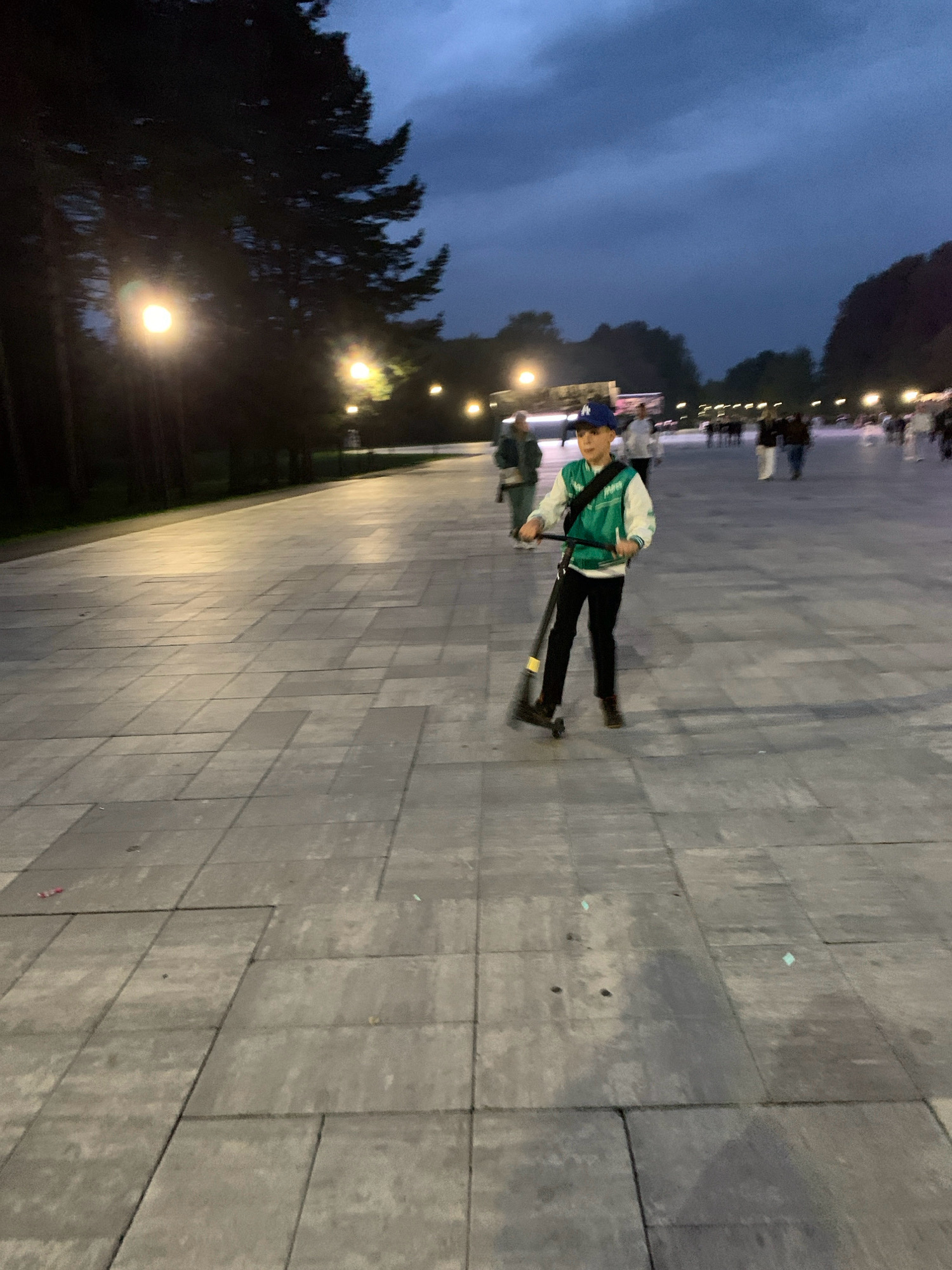 A young person wearing a blue baseball cap, a teal and white jacket, and black pants is riding a black scooter on a paved plaza at dusk. The plaza is made of large, grey rectangular tiles. Streetlights are visible in the background, casting yellow light on the ground and illuminating the scene. Trees are present on the left side of the image, silhouetted against the twilight sky. Other people are walking in the background, some blurred due to motion or distance. The sky is a deep blue, indicating the transition from day to night. The location is identified as a public plaza in Prokopyevsk, Russia, based on external information.