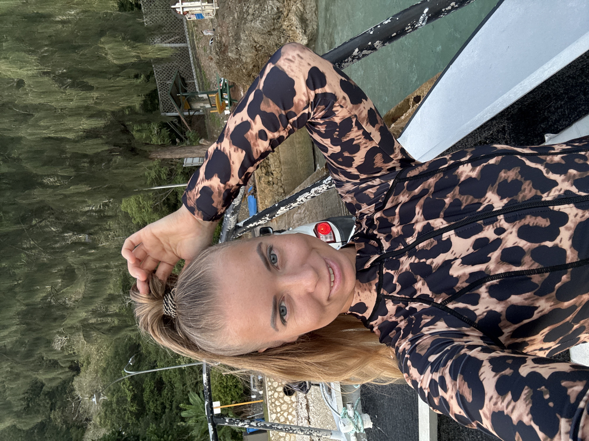 A blonde woman with blue eyes smiles at the camera, adjusting her high ponytail. She is wearing a long-sleeved leopard print rashguard. She is on a boat, with the ocean and a rocky, vegetated shoreline in the background. A car is visible parked near the shore. The lighting suggests it is either early morning or late afternoon. The location is Christmas Island, an Australian territory in the Indian Ocean, approximately 350 kilometers south of Java and 1,300 kilometers northwest of the Australian mainland.