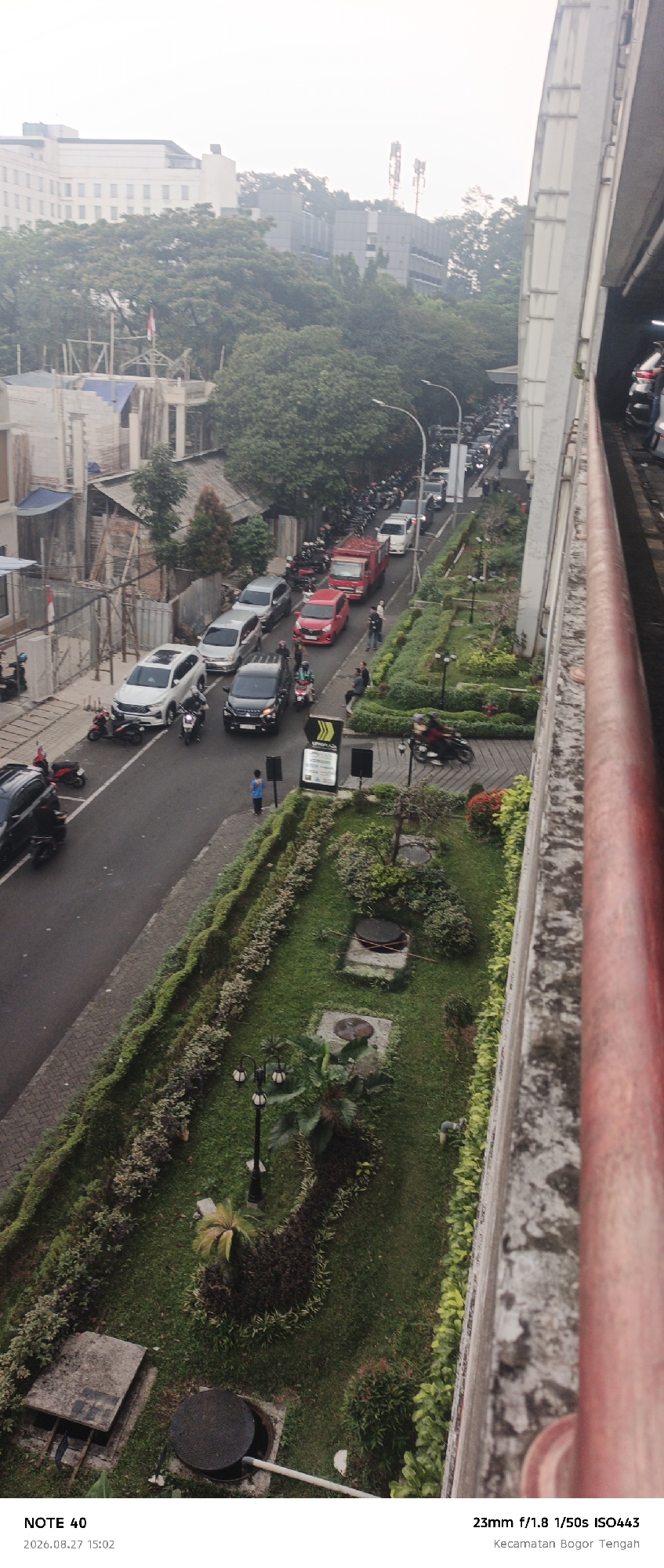The image captures a street scene in Kecamatan Bogor Tengah, Bogor, Indonesia. A traffic jam is visible on a moderately sloped road, with numerous cars and motorcycles lining up. A red truck is positioned in the middle of the traffic flow. On the right side of the road, there is a landscaped area with manicured bushes, grass, and trees. Two lampposts are present in the landscaped section. In the foreground on the left, a white SUV and a black SUV are stopped on the road, with several motorcycles parked on the side. A construction site with scaffolding is visible in the background on the left, with a flag prominently displayed on top of one of the structures. The sky is overcast. The bottom of the image contains text overlay, indicating "NOTE 40 2026.08.27 15:02" and "23mm f/1.8 1/50s ISO443 Kecamatan Bogor Tengah".