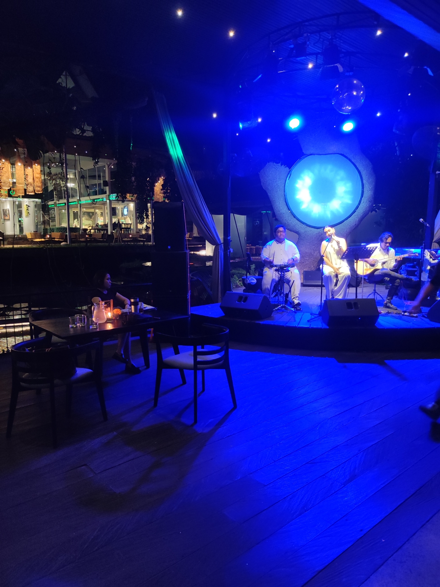 The image depicts a live music performance at a venue, possibly a restaurant or lounge, in Ubud District, Indonesia.
**Setting:** The scene is set at night. The venue appears to be outdoors, with a wooden floor and a covered stage area illuminated by blue stage lighting and a disco ball. In the background, a building with glass walls and greenery can be seen, suggesting a commercial area.
**People:** Three musicians are performing on stage. One is seated and playing a percussion instrument; another is singing and standing; and the third is playing an acoustic guitar and sitting. A woman is seated at a table in the foreground. A partially visible person is standing on the stage to the right of the musicians.
**Objects:** There are tables and chairs in the foreground, with drinks and candles on the table where the woman is seated. Stage equipment like speakers and microphones are visible. A large, circular light feature or decoration is behind the musicians. A white curtain is draped to the left of the stage.
**Actions:** The musicians are performing live music. The woman at the table is watching the performance.
**Interactions:** The musicians are interacting with each other as they perform. The woman is observing the performance.
**Environment:** The overall atmosphere is one of entertainment and relaxation. The blue lighting creates a cool and somewhat mysterious mood. The location in Ubud District, Indonesia, suggests a tropical or semi-tropical environment.