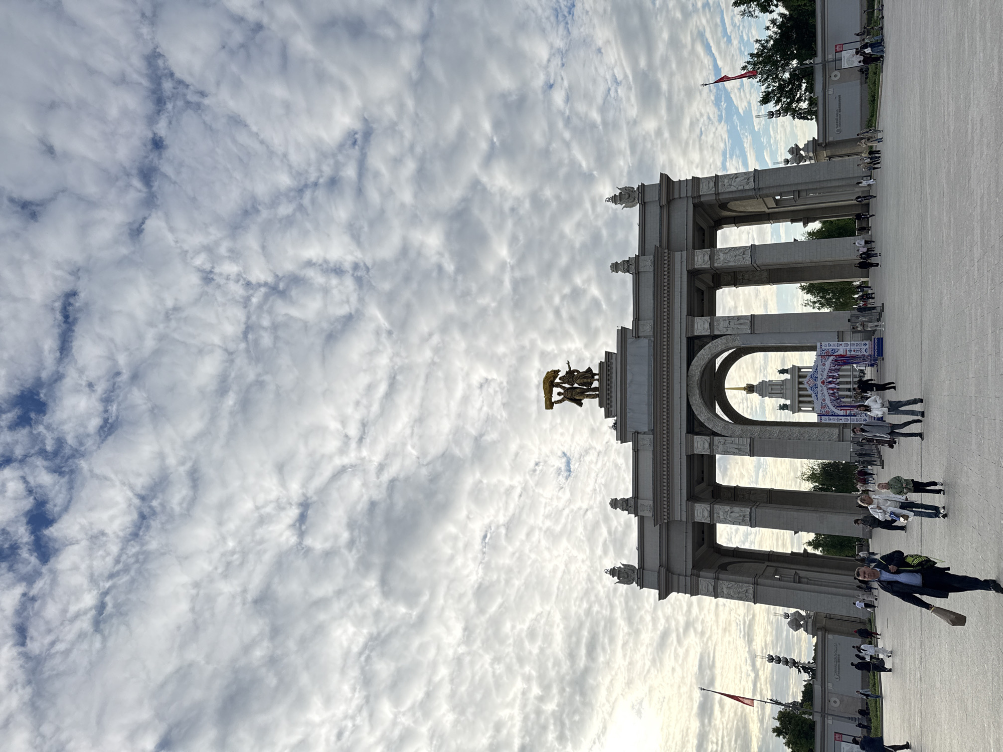 An outdoor, eye-level shot captures a grand architectural archway dominating a spacious public square in Moscow, Russia. The sky overhead is a prominent feature, filled with extensive, textured altocumulus clouds, suggesting an overcast yet bright late afternoon or early evening.
The monumental archway, constructed from light-colored stone or concrete, boasts multiple arched passages and ornate decorative details. A significant golden sculpture is positioned atop the central part of the arch, depicting stylized figures holding an emblem, consistent with Soviet-era monumental art often found at locations like VDNH. In the background, visible through one of the arches, stands another distinctive building with a prominent spired dome, further confirming the iconic setting of VDNH (Vystavka Dostizheniy Narodnogo Khozyaystva).
Numerous individuals are seen walking across the paved square, some passing through the archways, others milling about the foreground and background. Their relaxed movement suggests visitors enjoying a public park or exhibition ground. A red flag is visible on a pole to the left of the arch.
Underneath one of the central arches, several banners or signs are displayed. A prominent banner features Cyrillic text, including what appears to be "ВЫСТАВКА" (Exhibition) and "ВДНХ" (VDNH), clearly identifying an event or location. The overall ambiance is calm and leisurely, illuminated by the soft, diffuse light filtering through the clouds.