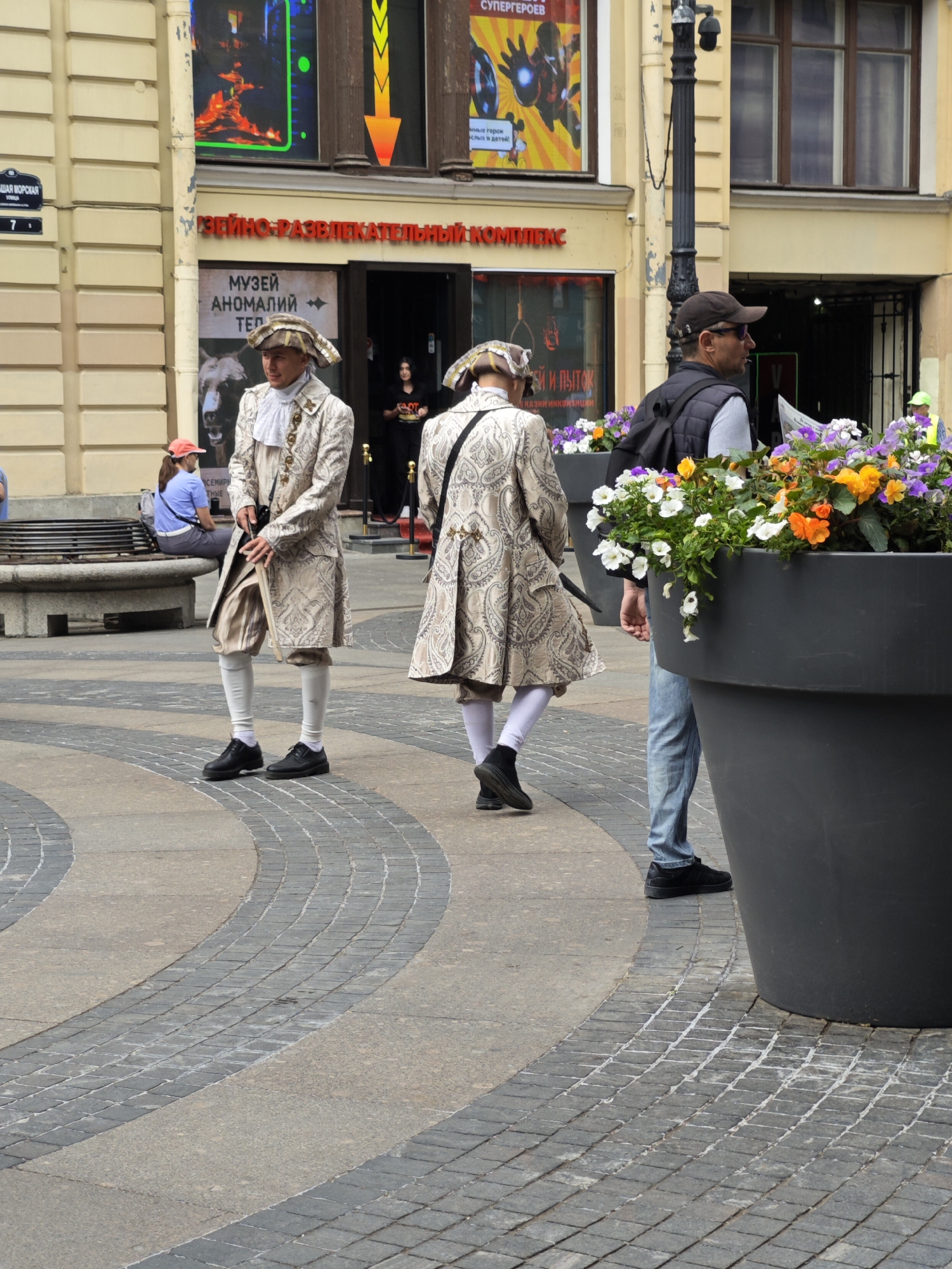 The image depicts a street scene in Saint Petersburg, Russia, likely on Nevsky Prospekt, a famous avenue known for its historical architecture and bustling atmosphere. In the foreground, two individuals are dressed in elaborate 18th-century-style costumes, complete with powdered wigs and ornate jackets. They appear to be performers or re-enactors, possibly part of a historical tour or event. One performer is standing with his back to the camera, while the other faces slightly away, holding a prop.
Behind them, a man in modern casual wear, a dark vest, and a baseball cap, stands next to a large, dark grey planter filled with colorful flowers. He is looking towards the performers, possibly as a spectator. Further in the background, a woman in a blue shirt and red cap is seated on a bench, observing the scene. The architecture visible includes the yellow facade of a building with large windows, and a red and yellow banner advertising an "ENTERTAINMENT COMPLEX" and a "MUSEUM OF ANOMALIES" is prominently displayed above an entrance. The ground is paved with cobblestones in a decorative pattern. The overall atmosphere suggests a lively tourist area.