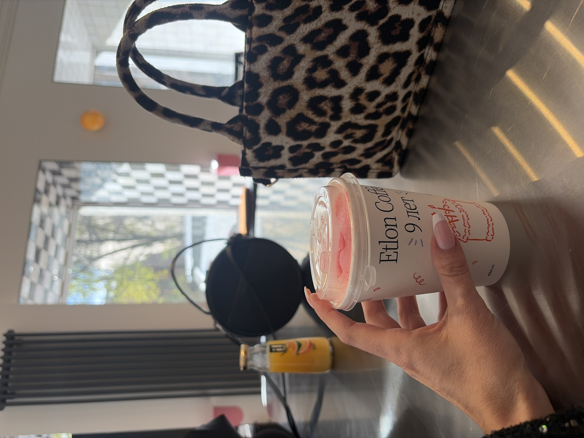 The scene captures an indoor setting, likely within a residence or studio, with a hand holding a disposable cup filled with a pink substance, presumably a beverage. The cup bears the text "Etlon Coffee," and the Cyrillic words "9 лет," indicating a coffee brand named Etlon, celebrating a 9-year anniversary. The context suggests the city of Saint Petersburg, Russia, due to the Russian text on the cup and the window view.
In the background, a leopard-print tote bag rests on a metallic surface, catching the light from the window. Behind the cup and hand, a black microphone with a pop filter is positioned, implying the presence of recording or audio equipment. A bottle of orange juice is partially visible behind the microphone.
The light and shadows across the metallic surface suggest the time of day is likely morning or late afternoon, with sunlight streaming in through the window. The window itself reveals a glimpse of the outdoors with trees, suggesting a residential or urban environment.
The focus is primarily on the hand holding the cup and the surrounding elements, while the background elements are less sharp, creating depth in the image. The overall setting suggests a casual and potentially creative environment.