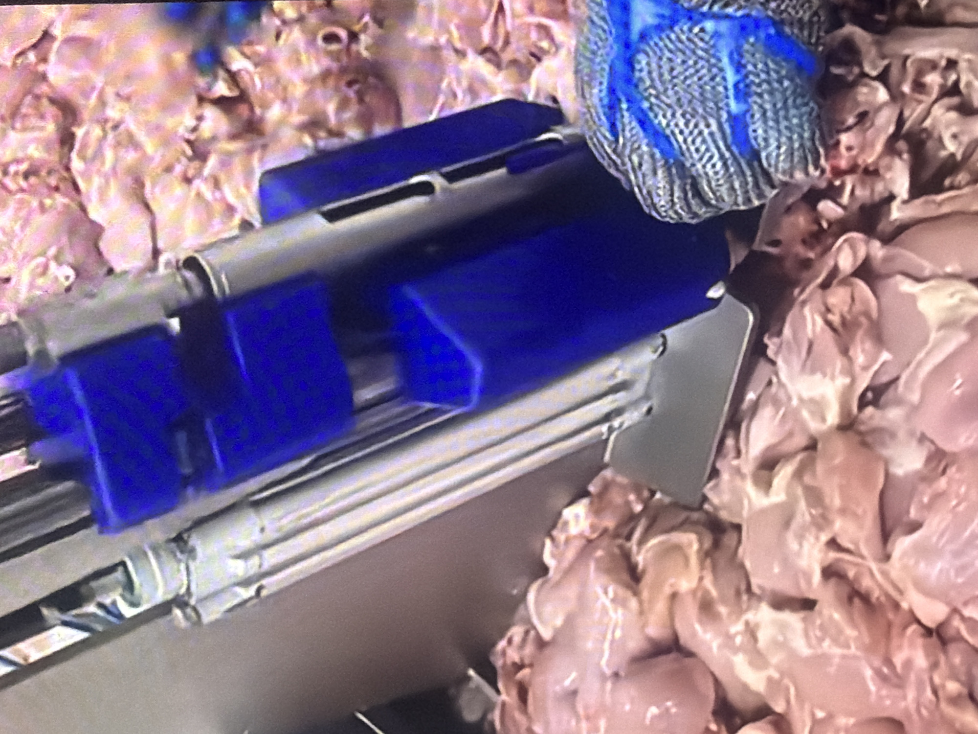 The image shows a close-up view of raw chicken pieces in what appears to be a retail or processing environment. A piece of metal equipment, possibly a cutting or processing tool, with blue plastic components is positioned over the chicken. The chicken is pale pink and appears to be boneless thighs. The background shows more chicken in a larger container, with a pinkish hue. The overall setting suggests a food preparation or sale area. There are no people visible in the image.