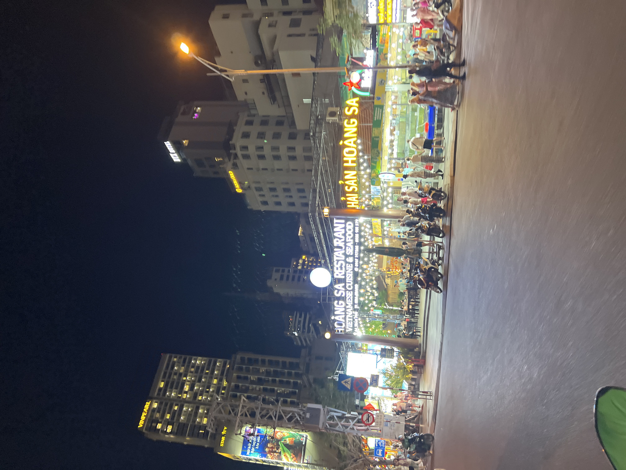 The image depicts a nighttime scene in Nha Trang, Vietnam, showcasing a busy street with restaurants and hotels. "Hải Sản Hoàng Sa" restaurant is prominently featured with bright signage. People are seen walking and dining. Motorbikes are parked along the street. The "Azura Gold" hotel is visible in the background, along with the "Vinpearl" and "DIX HOTEL" brands on buildings. Streetlights illuminate the scene.