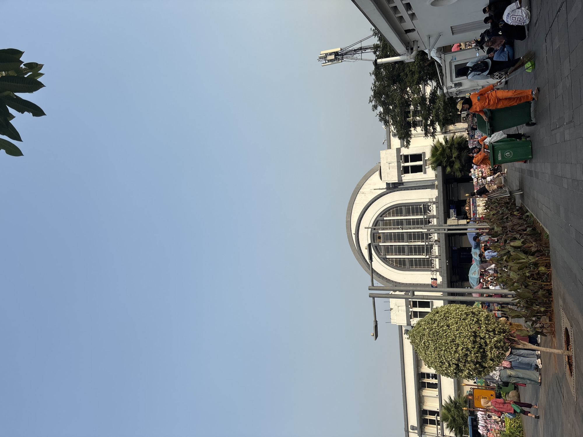 The image captures a scene in front of the historic Jakarta Kota train station in West Jakarta, Indonesia. The sky is clear and blue. In the foreground, a paved walkway is lined with lush green plants and a single, neatly trimmed tree. Several individuals are present, including a group of three sanitation workers in bright orange uniforms. One worker is pushing a green trash bin, another is sweeping the ground with a broom, and a third is standing nearby. Other people are seen walking, sitting, or standing around, some near market stalls or a building. The architecture of the train station, characterized by its large arched windows and white facade, is visible in the background. A telecommunications tower stands to the right of the station. The overall atmosphere appears to be a busy urban environment during daylight.