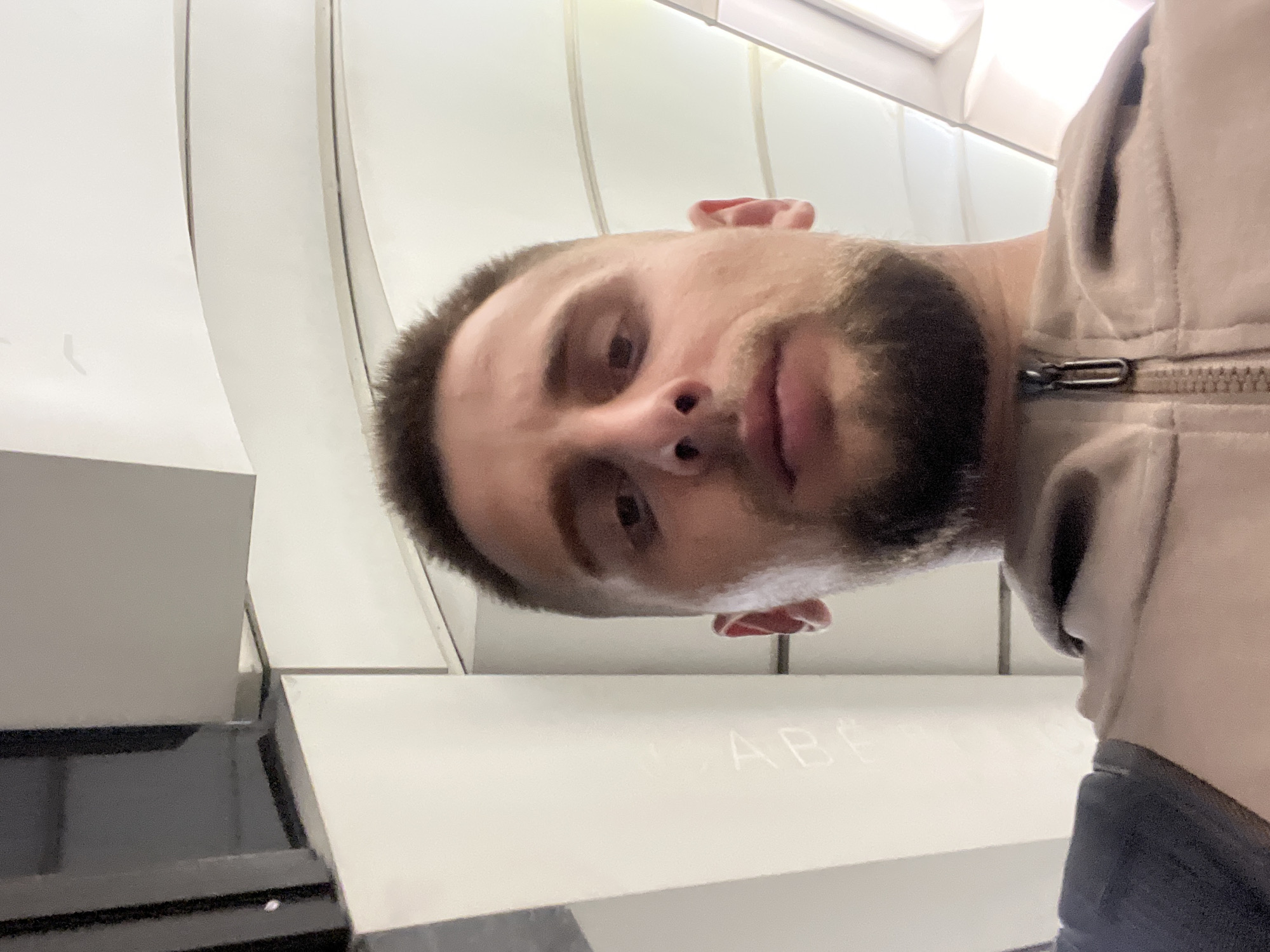 A low-angle, close-up, first-person perspective selfie captures a man with short brown hair and a beard. He is wearing a light-colored hooded jacket with a zipper. The background consists of abstract architectural elements, appearing to be interior surfaces with clean lines and curves, possibly a modern building or transportation hub. The lighting suggests an indoor environment with artificial light sources. No specific location in Moscow, Russia, can be identified from the provided image.