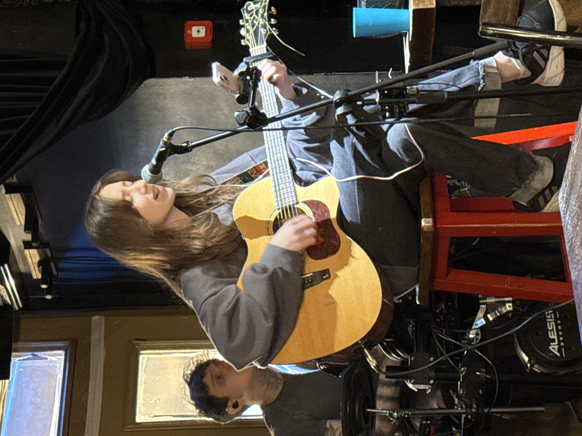 An indoor, portrait-oriented photograph captures a live musical performance. The main subject is a young woman, seated and actively singing into a microphone while playing an acoustic guitar. She has long, straight brown hair and is dressed in a gray long-sleeved top and ripped blue jeans, paired with black and white sneakers. Her mouth is open in song, and her hands are engaged with the light-wood-toned acoustic guitar, which features a darker pickguard and an ornate headstock with a capo attached.
The setting appears to be a small performance venue or bar, characterized by dim lighting and stage equipment. In the background, dark curtains are draped, and a red fire alarm call point is mounted on a wall. A drum set, bearing the "ALESIS DRUMS" logo, is partially visible behind the performer, along with various microphone stands. A white smartphone is attached to one of the mic stands, seemingly recording the performance.
A second person, a man with dark, curly hair and an ear gauge, wearing a dark t-shirt, is partially visible in the lower right background, looking towards the performer. The scene suggests an intimate musical event taking place in Kazan, Russia, likely in the evening.