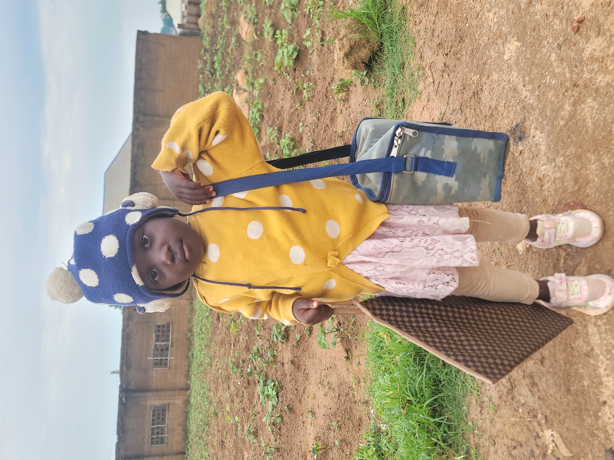 A young child, likely female, stands in an outdoor, rural setting. She is wearing a yellow sweater with white polka dots, a light pink lace skirt, beige leggings, and pink sneakers. On her head, she has a blue knitted hat with white polka dots and two pom-poms. She is holding a flat, brown, checkered mat in her left hand and has a camouflage lunch bag with a blue strap slung over her right shoulder. The background shows a dirt path, sparse vegetation, and a low, brown building with barred windows, consistent with a school or community building. The sky is overcast. The scene appears to be located in Barakin Akawo, Nigeria.