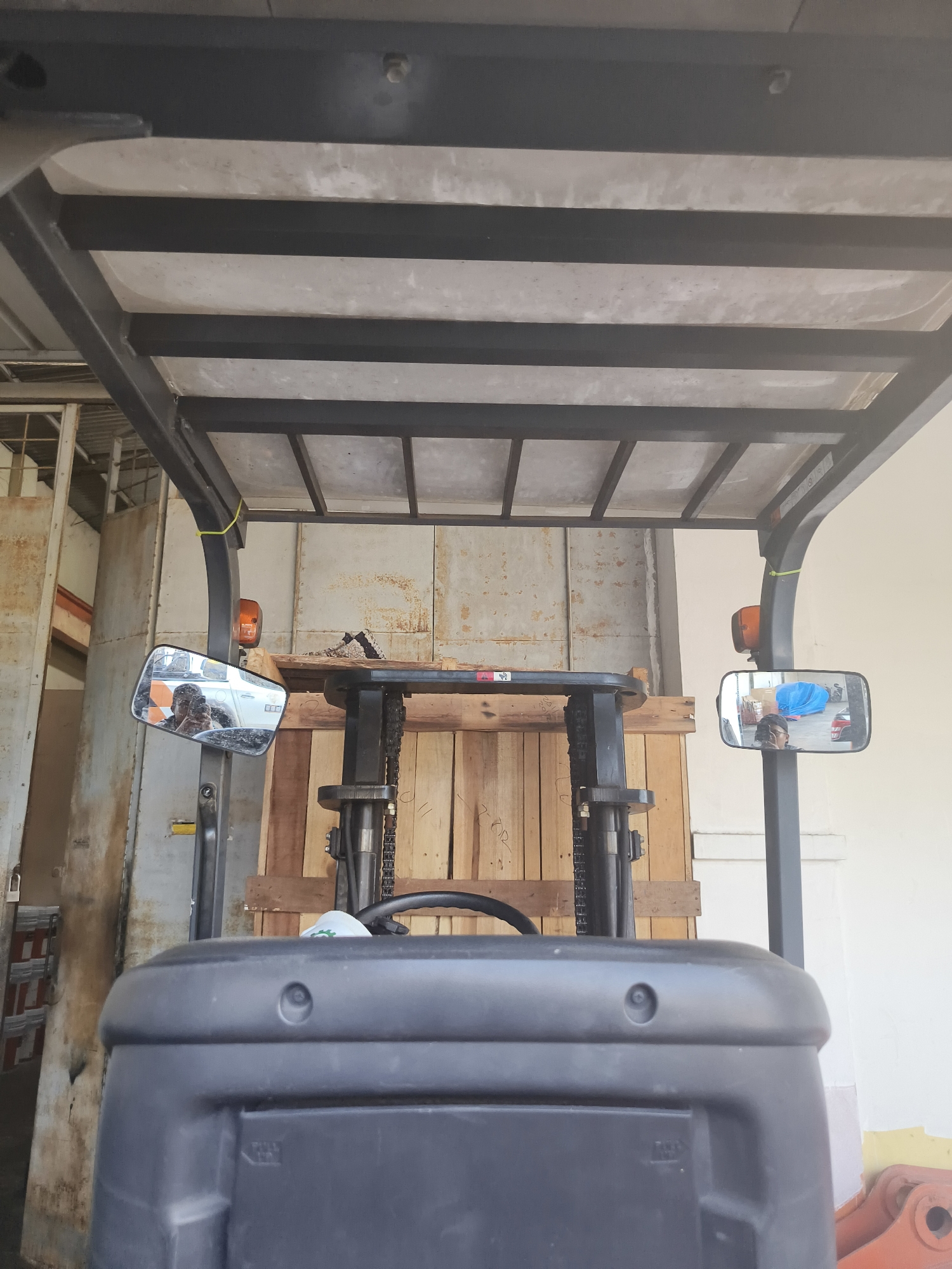 The image shows the interior view from the driver's seat of a forklift. The forklift has a protective canopy overhead, supported by black metal frames. On either side of the driver's area, there are rearview mirrors. The left mirror reflects a person wearing a dark shirt and a vehicle. The right mirror reflects a person wearing glasses and a dark shirt, with a blue object and a vehicle in the background. In front of the driver's seat, there is a steering wheel and a wooden pallet is visible behind it. The environment appears to be a warehouse or industrial setting, with metal sheeting and wooden structures visible in the background. There is no specific indication of the location in Pangkalpinang, Indonesia.