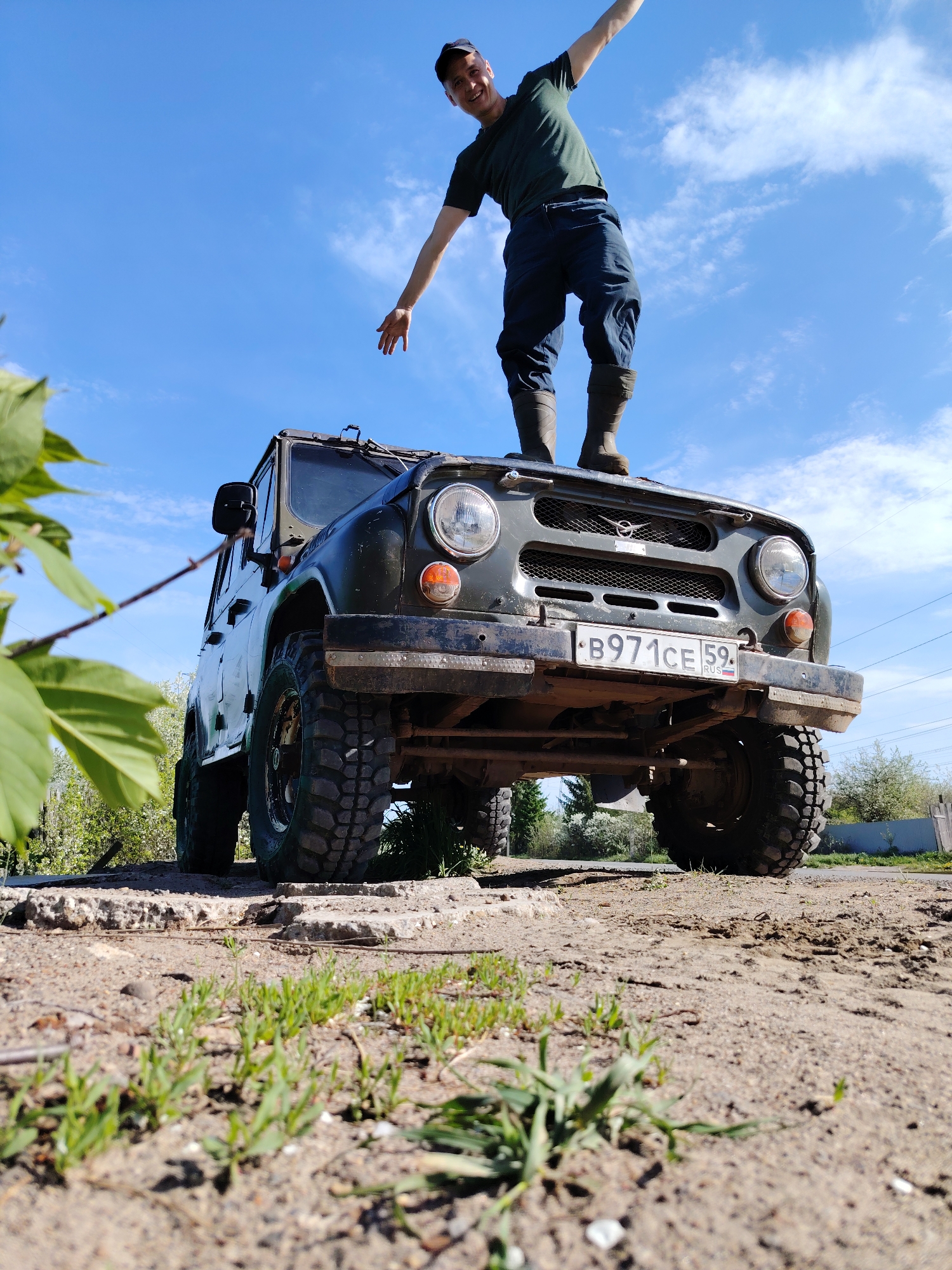 A man is standing on the hood of a dark-green UAZ-469 off-road vehicle. The license plate reads "B971CE 59" which indicates it is registered in the Perm region of Russia. The man is wearing a dark-green short-sleeved shirt, dark pants, and dark boots. His arms are outstretched, one pointing downward and the other raised in the air. The vehicle is parked on a dirt surface mixed with concrete debris and small plants. In the background, there are trees and a residential fence. The sky is blue with scattered clouds. Based on the license plate, the photo was most likely taken in the Perm region of Russia. The location is confirmed to be Chaikovsky, Russia.