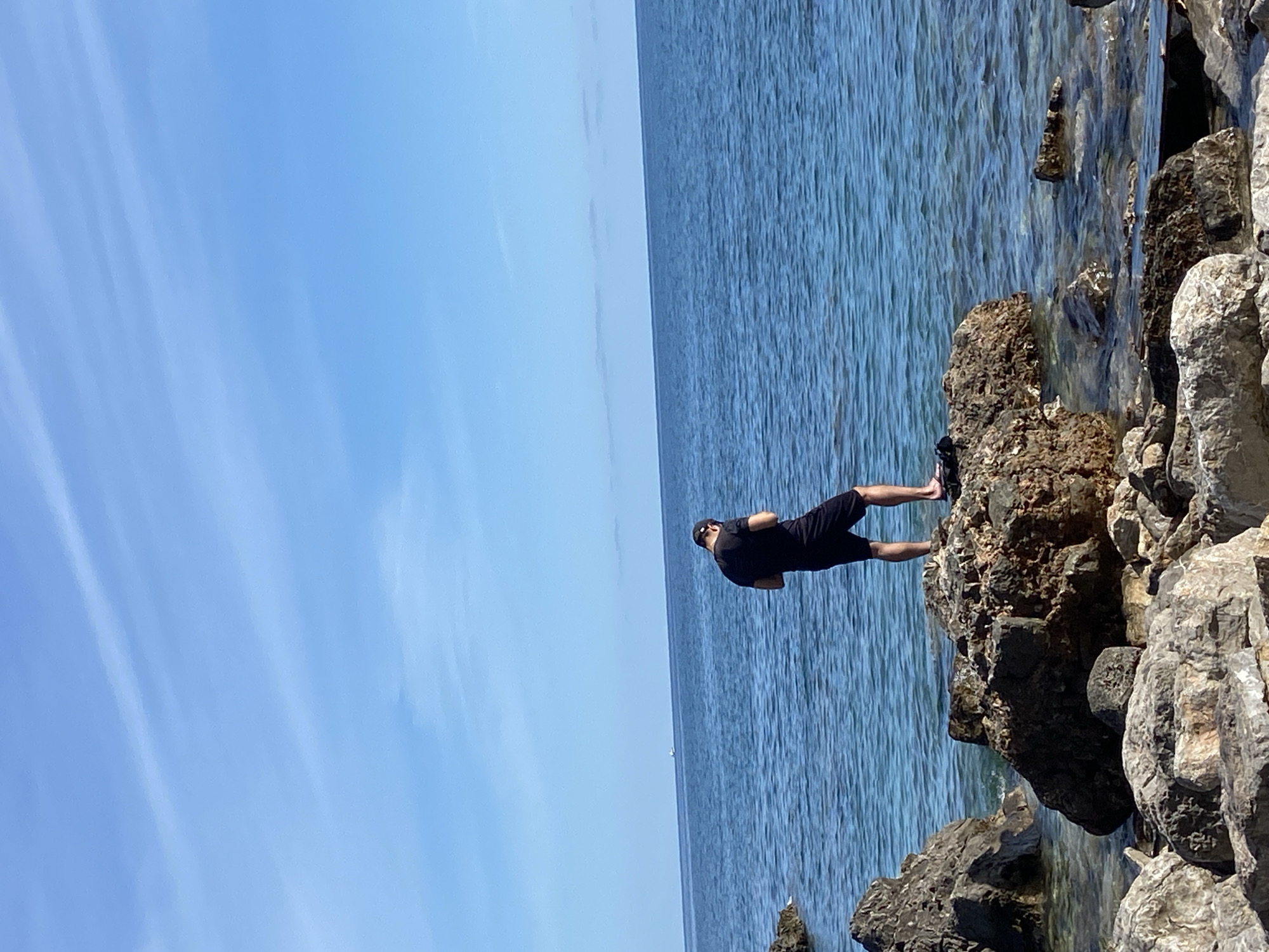 A medium shot depicts a man standing on a rocky outcrop in Prijevor, Montenegro, against the backdrop of the Adriatic Sea and a clear, light blue sky. The man, who is wearing black shorts and a black T-shirt, appears to be gazing out at the water. The surrounding environment is characterized by jagged rocks that meet the sea, suggesting a coastal location.