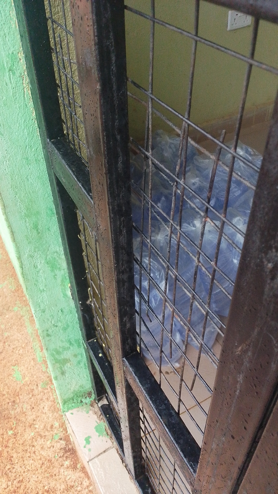 The image shows a close-up of a black metal gate with vertical and horizontal bars, through which stacks of blue-wrapped items are visible. The gate appears to be on an exterior wall painted green, with a concrete floor at the base. The lighting suggests it may be daytime, possibly overcast due to the diffused light. The scene is outdoors, with what appears to be loose soil and some tiling visible near the base of the gate. The overall condition of the gate and surrounding elements suggests an older, possibly weathered, environment.