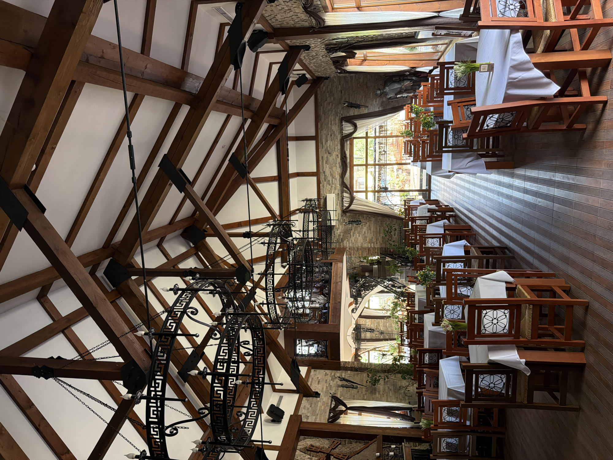 This image depicts the interior of a restaurant, likely located in Gorna Malina, Bulgaria. The space features a high, vaulted ceiling with exposed wooden beams and a white ceiling. Two large, ornate black chandeliers hang from the ceiling, each with multiple candle-like lights and intricate geometric detailing. The walls are constructed of stone or brick, with some sections featuring a textured, tiled pattern.
The main focus of the image is the dining area. Numerous tables, draped with white tablecloths, are set with wooden chairs. The chairs have woven seats and backs with decorative metal inserts. Small floral arrangements are visible on some of the tables. The flooring is made of polished wooden planks.
In the background, large windows allow natural light to enter the restaurant, and greenery can be seen outside. A staircase with an ornate railing leads to an upper level or mezzanine. A suit of armor stands near a wall in the background. The overall ambiance suggests a rustic yet elegant dining establishment. There are no people visible in the image.
