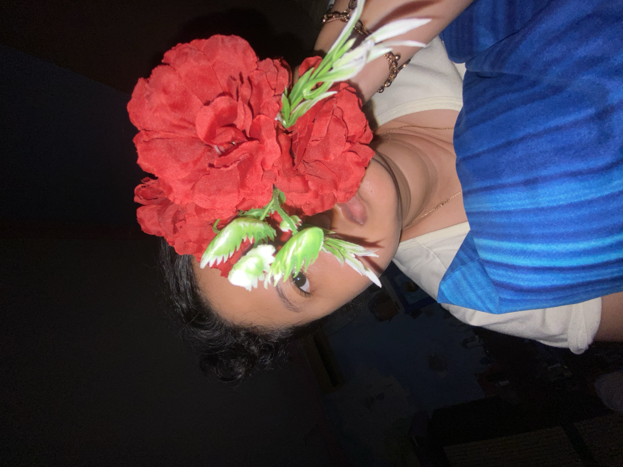 A close-up, low-light photograph shows a person holding a large, artificial red flower in front of their face, obscuring their left eye and nose. The person's visible right eye is dark brown. Their hair is dark and pulled back. They are wearing a cream-colored t-shirt and a blue garment with vertical stripes. A gold bracelet with small charms is visible on their right wrist. The background is dark and out of focus, with hints of a blue wall and possibly furniture. The scene appears to be indoors, in a room. No specific location in Gelam, Indonesia can be determined from the image.