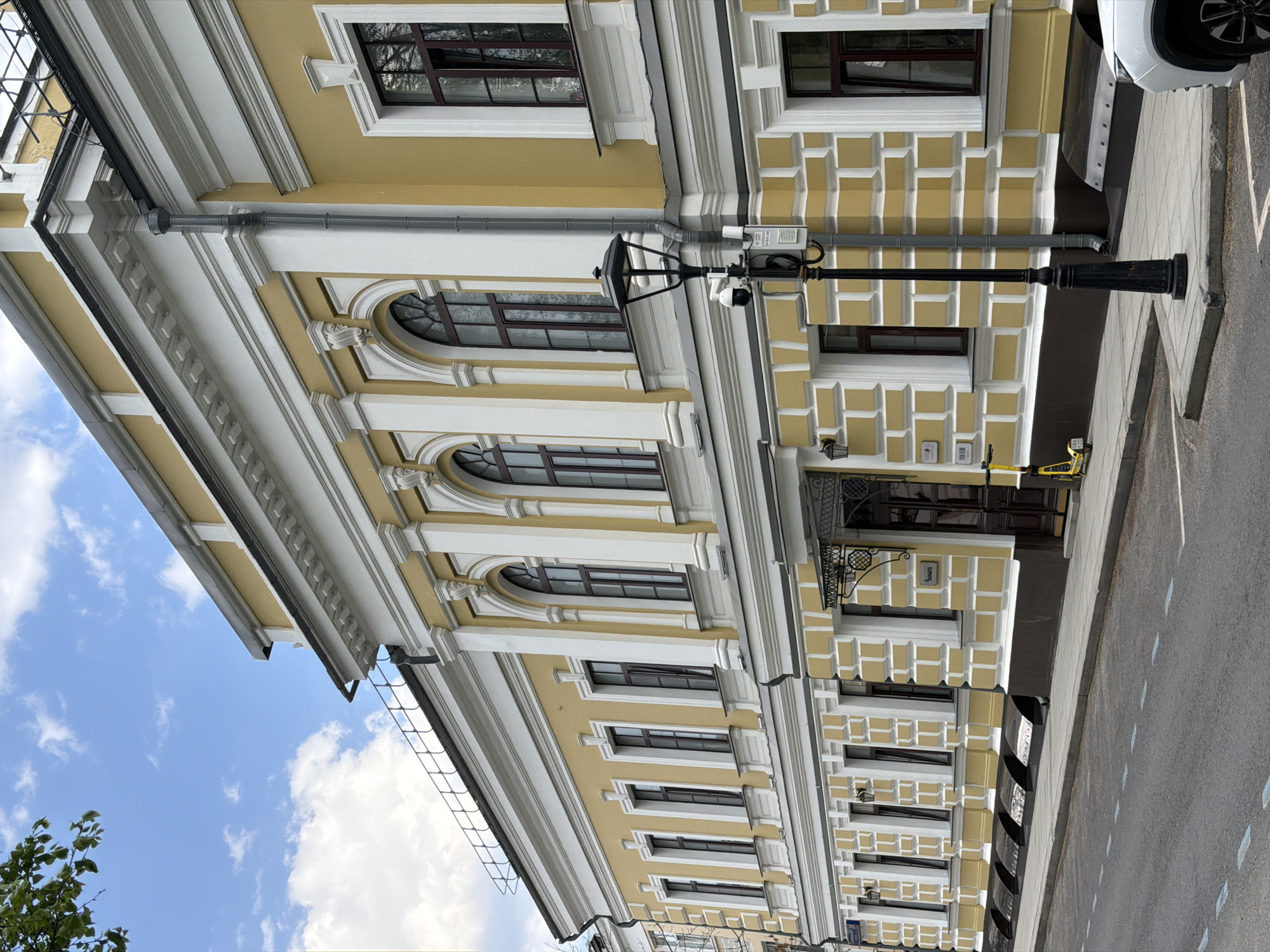 The image shows a building in Moscow, Russia. The building is yellow and white, with multiple stories and windows. The architecture is ornate. A black lamp post stands in front of the building, and there is a sidewalk and street visible. A car is partially visible at the edge of the image. The sky is blue with some white clouds.