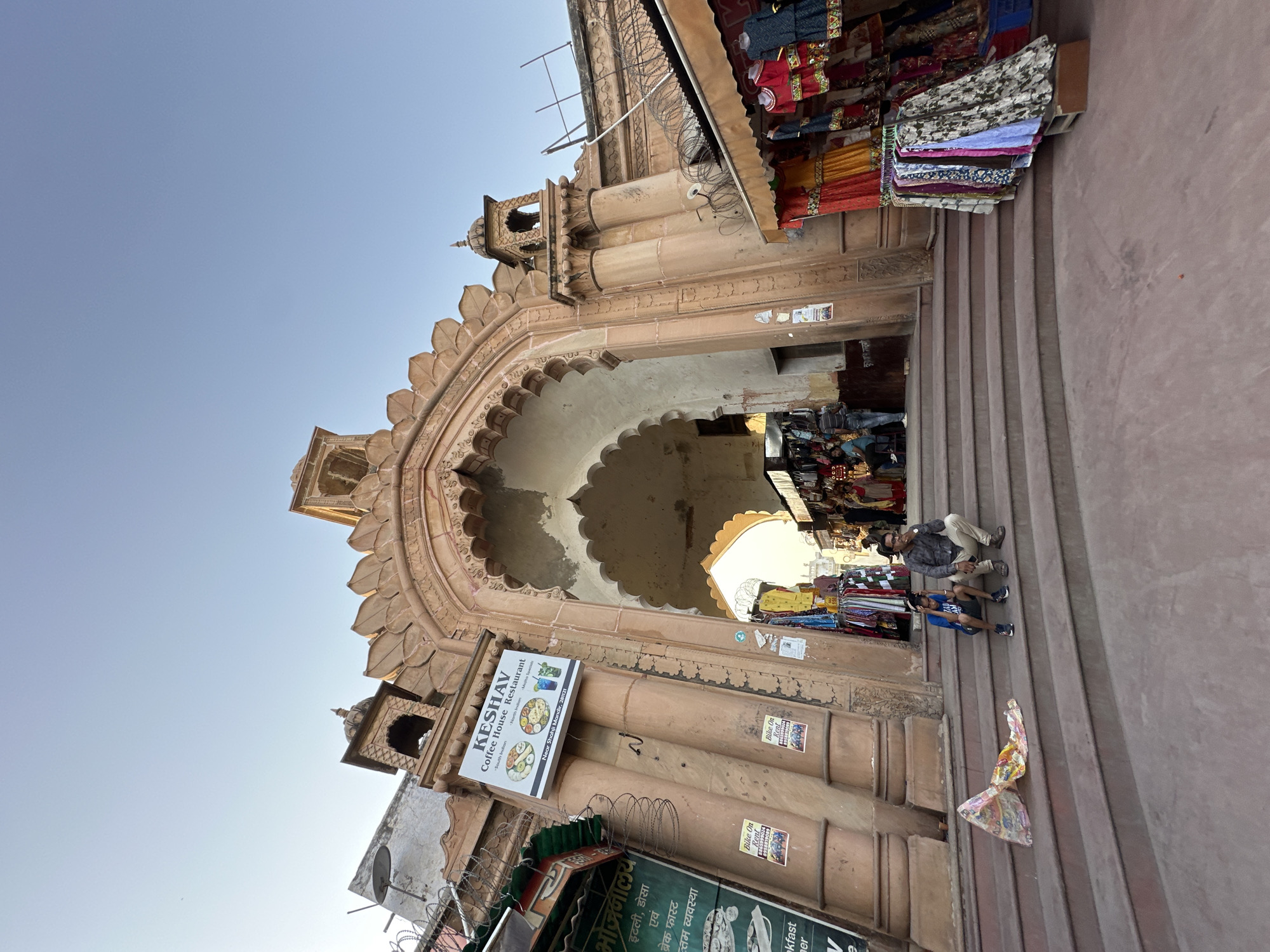 The image depicts an ornate arched gateway leading into a bustling market area in Vrindavan, India. The gateway is constructed from light brown stone, with intricate carvings and a scalloped archway. On the left side of the gateway, there are various signs and advertisements, including one for "KESHAV Coffee House Restaurant." To the right of the archway, merchandise, mainly colorful garments, is displayed on a small stall roof.
People are sitting on the steps leading up to the gateway. The sky is clear and light blue, indicating it is daytime. The overall atmosphere is vibrant and suggestive of a lively commercial area within a historic urban setting.