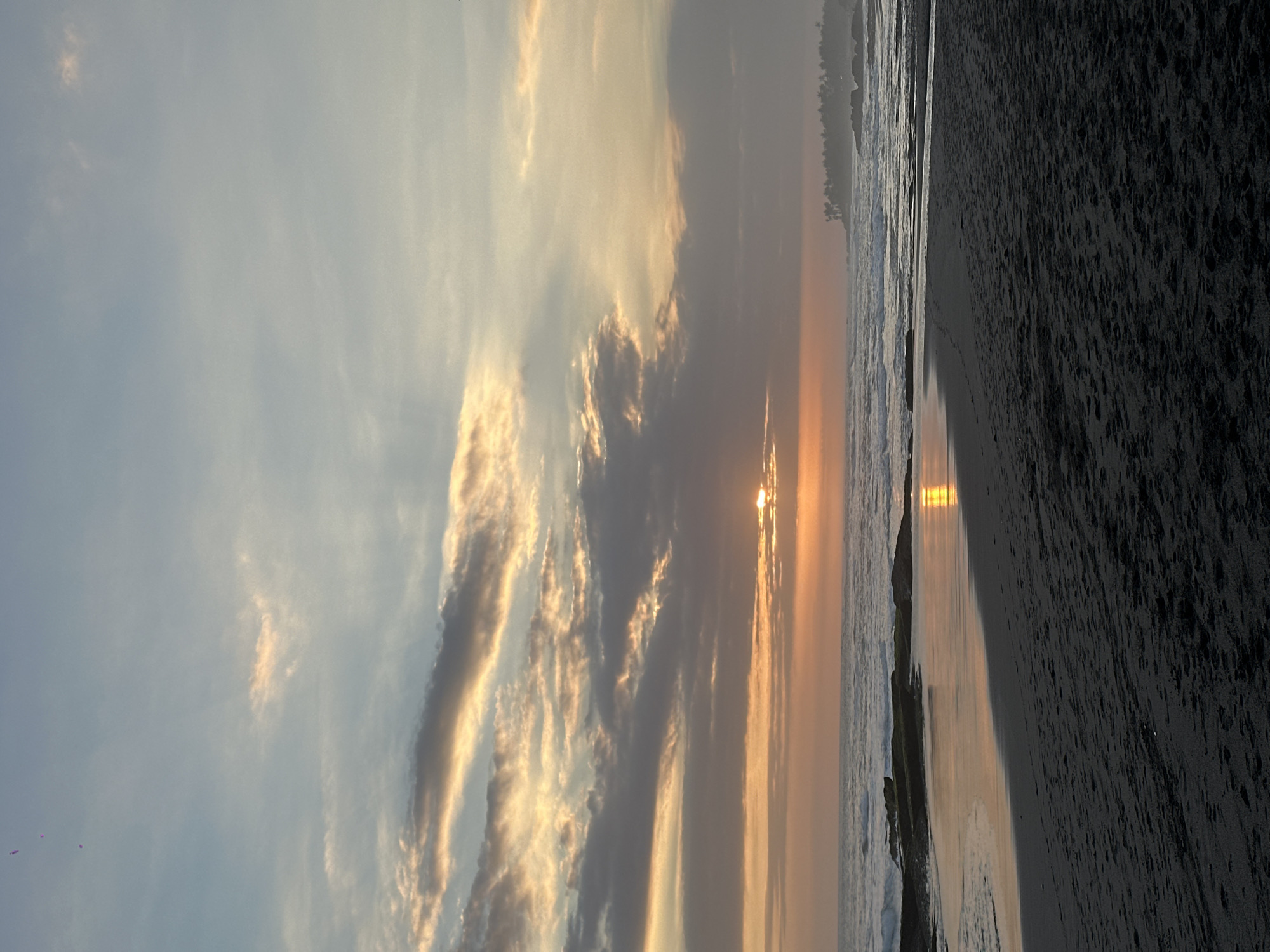 A vertical, medium shot captures a coastal scene at sunset. The sky, a gradient of pale blue and soft orange, is adorned with wispy clouds illuminated by the setting sun, which is partially obscured by clouds, casting a warm glow. The ocean, a deep blue, meets the shore with gentle waves. In the foreground, dark, textured sand forms a beach, transitioning to wet sand reflecting the sunset's colors. Rocks dot the shoreline, some partially submerged in the water. In the distance, a treeline with silhouetted palm trees suggests a tropical landscape. The image composition emphasizes the vastness of the sky and sea, with the beach occupying the lower portion.
