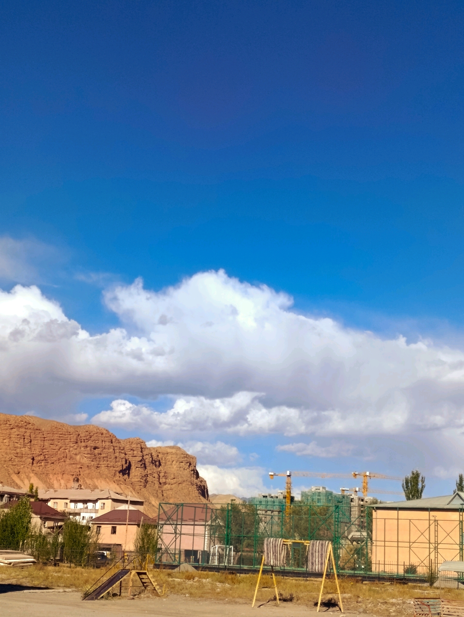The image depicts a scene in Naryn City, Kyrgyzstan, with a prominent backdrop of a large, reddish-brown rock formation. In the foreground, there is a playground area with a small slide and ladder structure. Beyond the playground, residential buildings are visible, some with a distinctly Soviet-era architectural style. Further in the distance, construction is underway, indicated by several tall yellow cranes and partially built structures covered in green netting. The sky is a bright blue with scattered white clouds. The overall impression is of a semi-urban environment bordering a natural landscape.