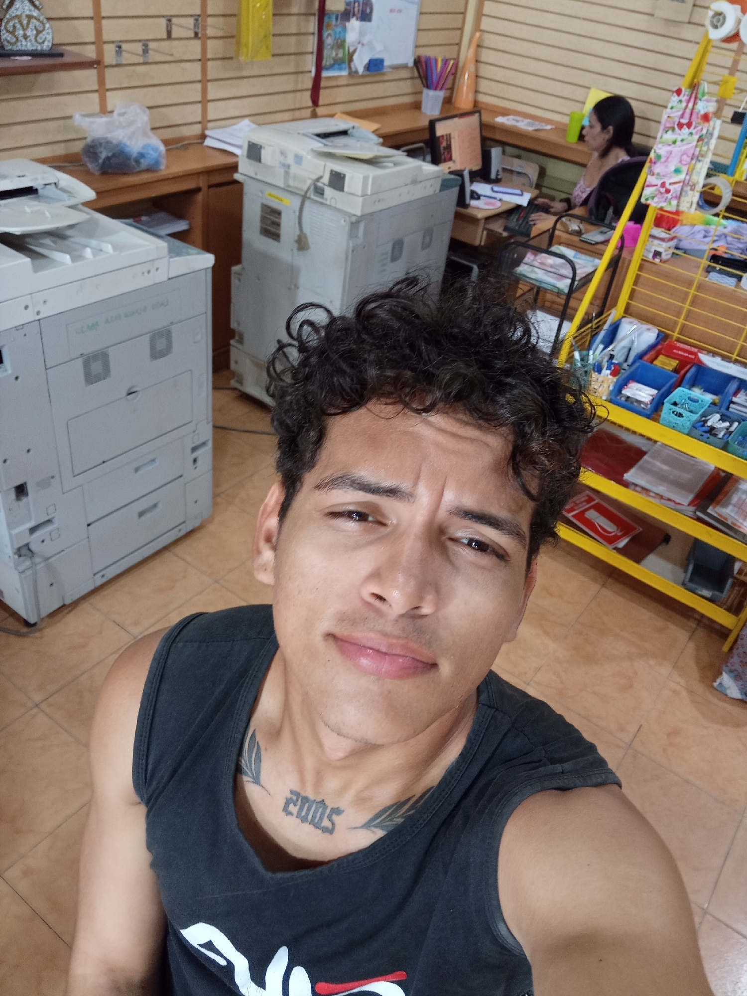 An indoor scene in Juan Griego, Venezuela, depicts a commercial establishment, likely a copy center or office supply store. In the immediate foreground, a young adult male with curly dark hair and light brown skin is taking a selfie, looking directly at the camera with a slight smile. He wears a dark grey sleeveless tank top, revealing a "2005" tattoo with flanking leaf designs on his lower neck/upper chest. His left shoulder and upper arm are visible.
The background reveals a functional workspace. To the left, a large, light grey multi-function copier stands prominently. Further back, on a wooden counter, a white plastic bag holds blue and black items. Another large, light grey copier is partially visible to the right of the first.
On the right side of the image, a woman with dark hair, pulled back, is seated at a wooden desk. She wears a light-colored, patterned top and is engaged with a computer monitor, keyboard, and what appears to be a desk phone or organizer. Adjacent to her desk is a yellow metal shelving unit with multiple tiers, displaying various items in blue and white bins (e.g., pens, office supplies) and some hanging merchandise bags.
The back wall features light wood paneling with slat-wall hooks. A decorative ornamental object rests on a shelf above the left copier. The floor is covered with light brown square tiles.