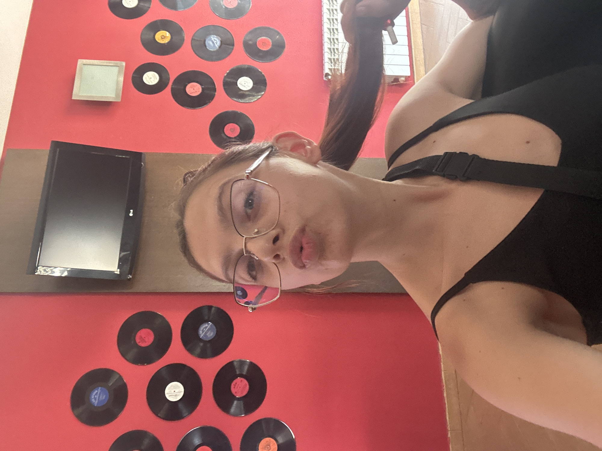 A young woman with glasses and brown hair pulled back in a ponytail is taking a selfie in front of a red wall decorated with vinyl records. She is wearing a black top. The background includes a small television and a white radiator. The location is the city of Tryavna, Bulgaria.